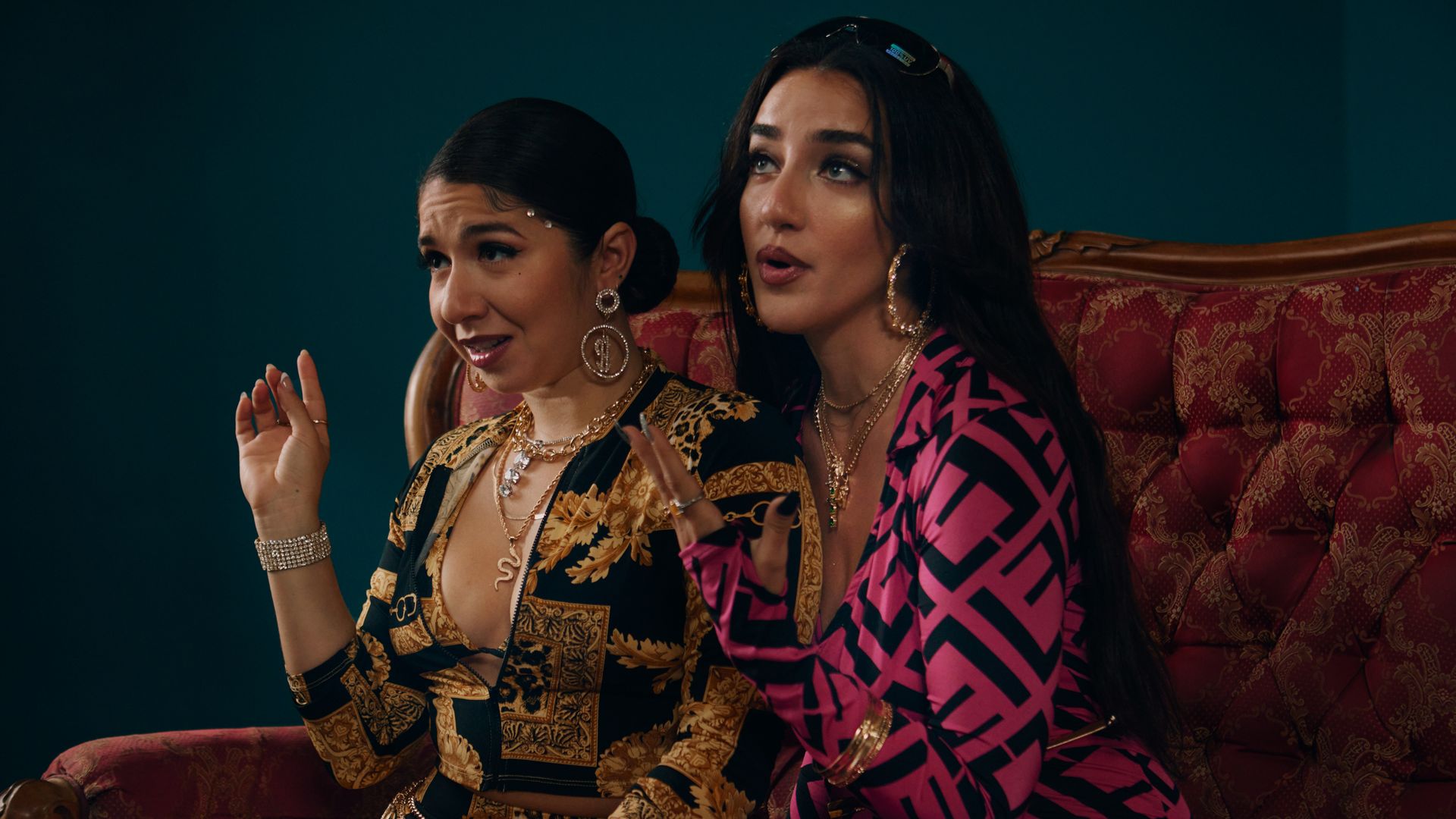 Two women in tight spandex outfits, heavy makeup and hoop earrings sit on a couch and look concerned.