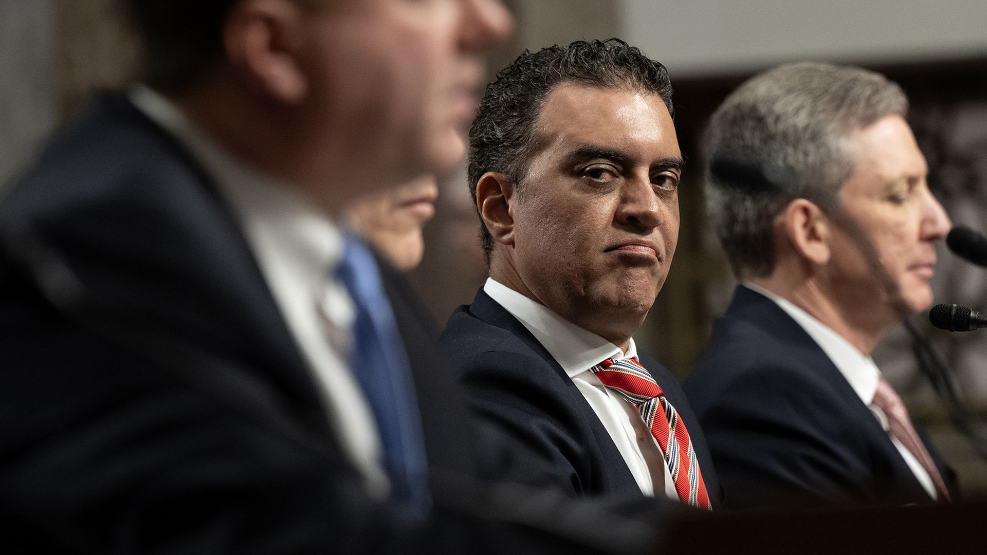 Four men in suits seated at a panel with microphones. One man in focus wearing a red striped tie and looking sideways with a serious expression.