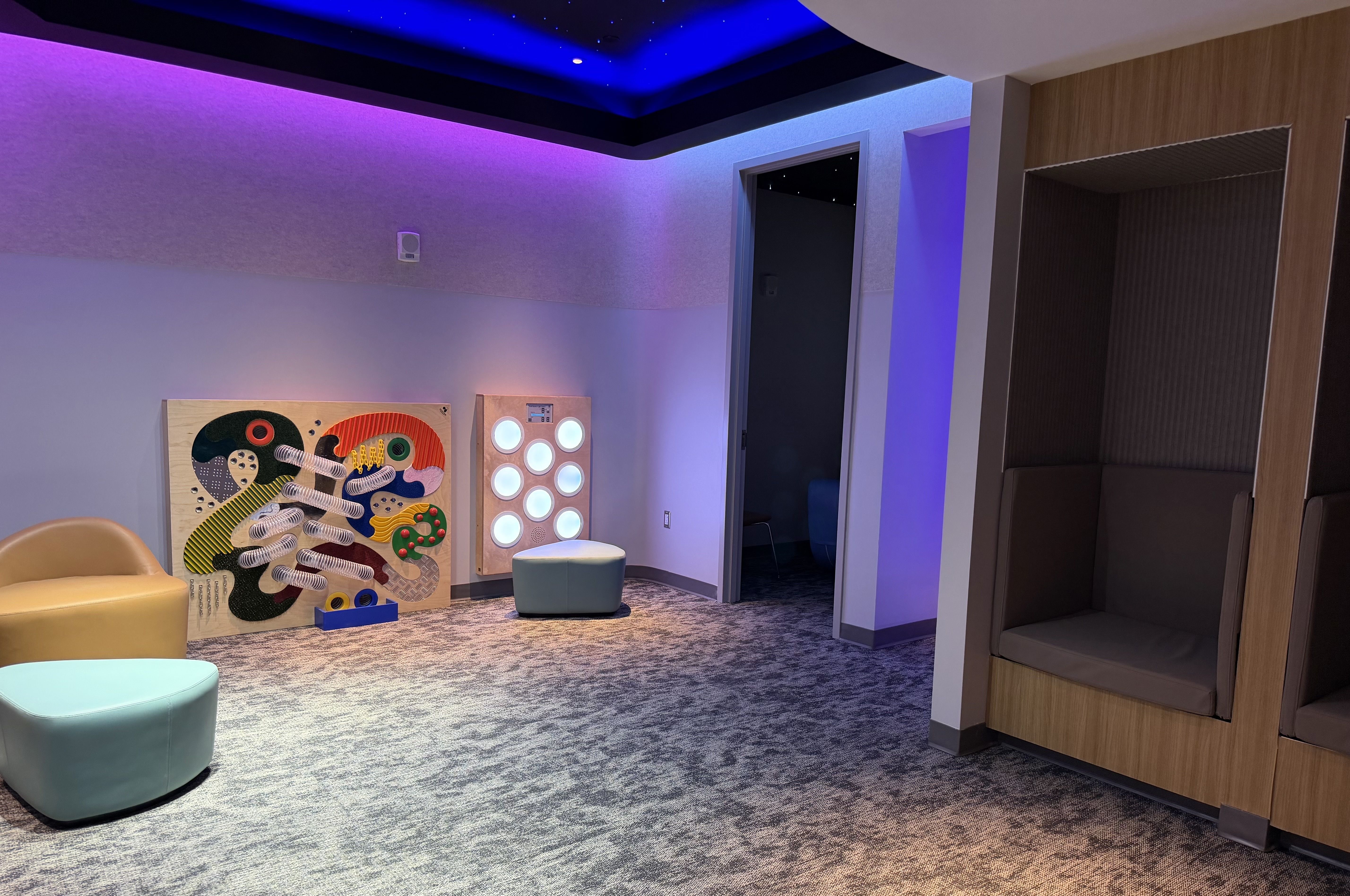 Modern lounge with purple and blue ceiling lighting, colorful abstract panels leaning against the wall, round teal ottomans, and wood-paneled seating along the right; doorway to a dark adjoining room.