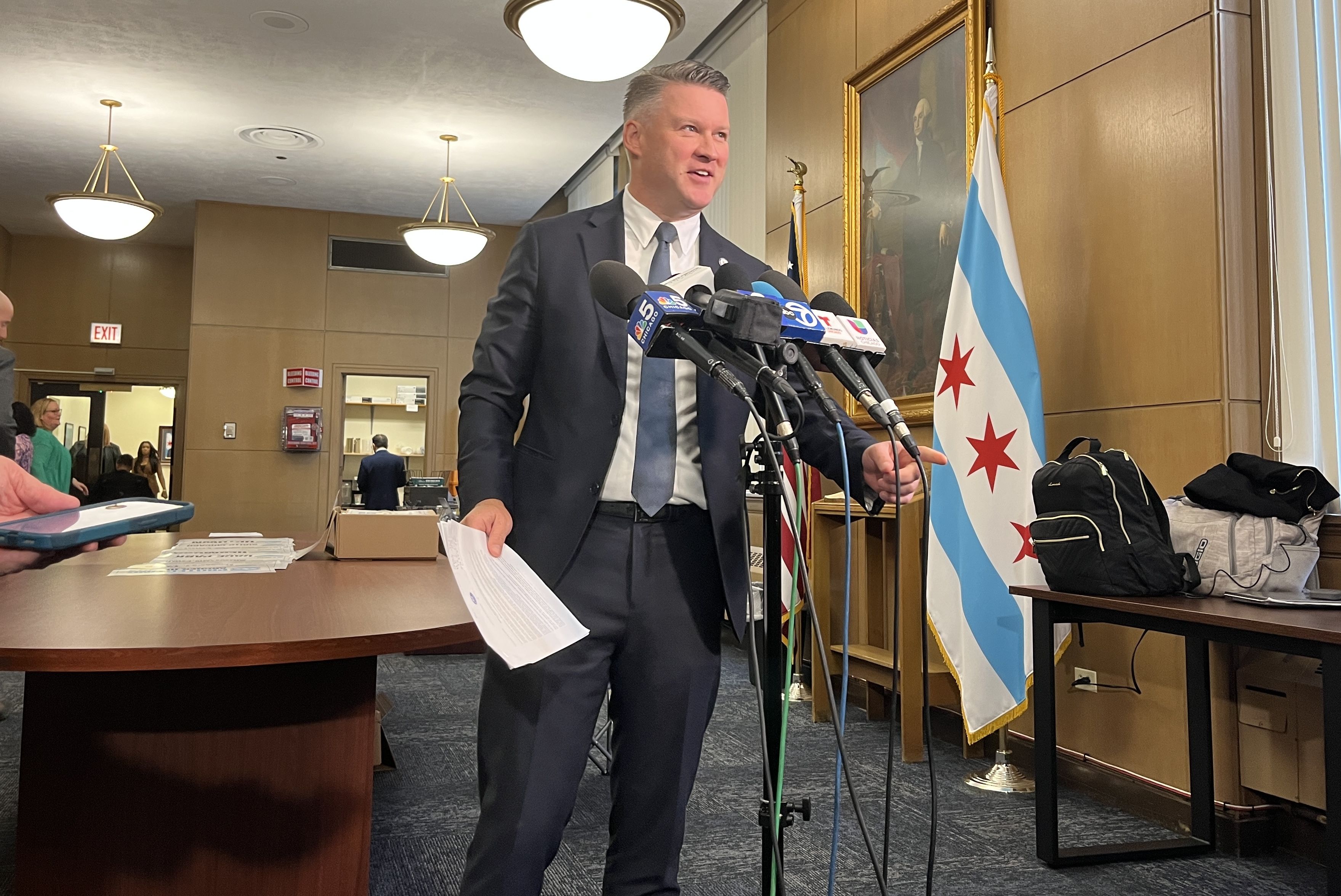Man in a dark suit speaking at multiple microphones in a room with wooden paneling, a Chicago flag, and a portrait behind him, holding papers and gesturing with his hand.