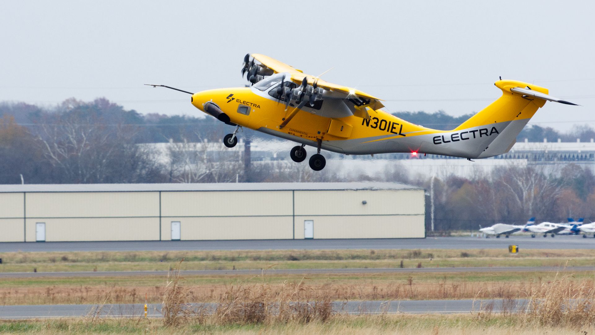 Yellow and white aircraft labeled Electra taking off from a runway on a cloudy day, with hangars and trees in the background.