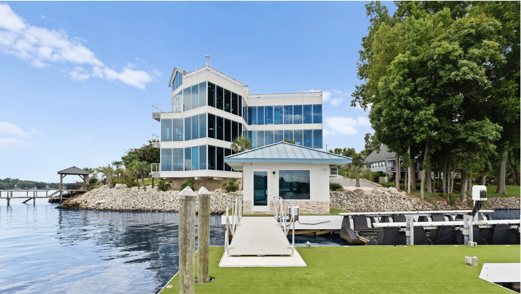 Modern multi-story glass building by a lake with rocky shoreline, green trees, a walkway leading to a small white dock house, and blue sky with scattered clouds.