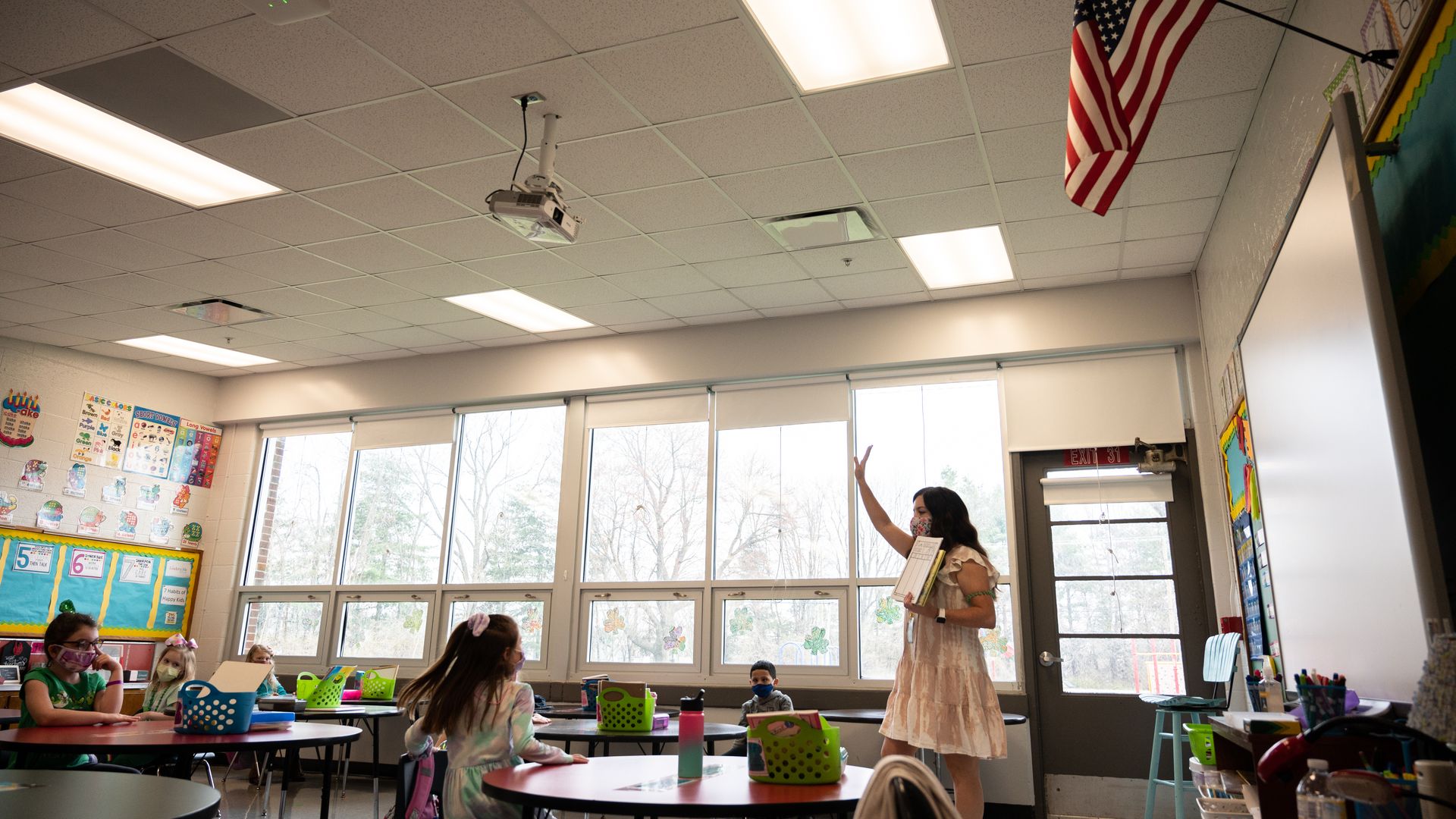 A teacher stands in front of a whiteboard raising her hand as she speaks to young students in a brightly lit classroom.
