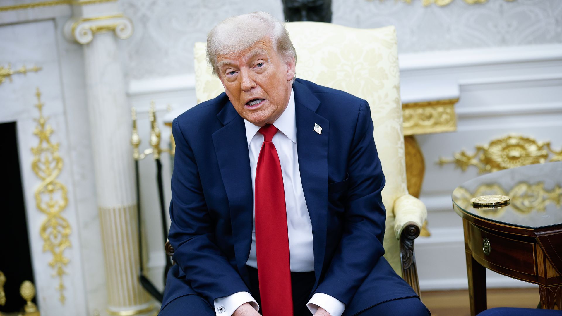 Man in dark blue suit, white shirt, and red tie sitting on cream chair with gold details, holding papers, speaking in an ornate room with gold and white decor.