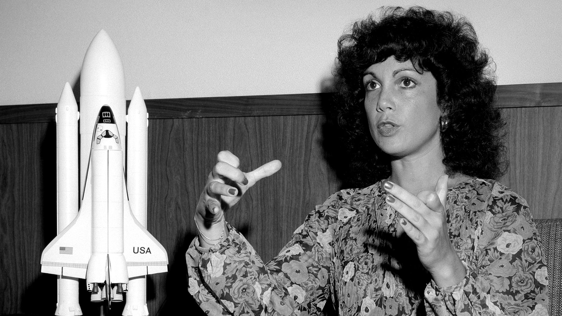 Black and white photo of woman with black curly hair talking with her hands beside a space shuttle.