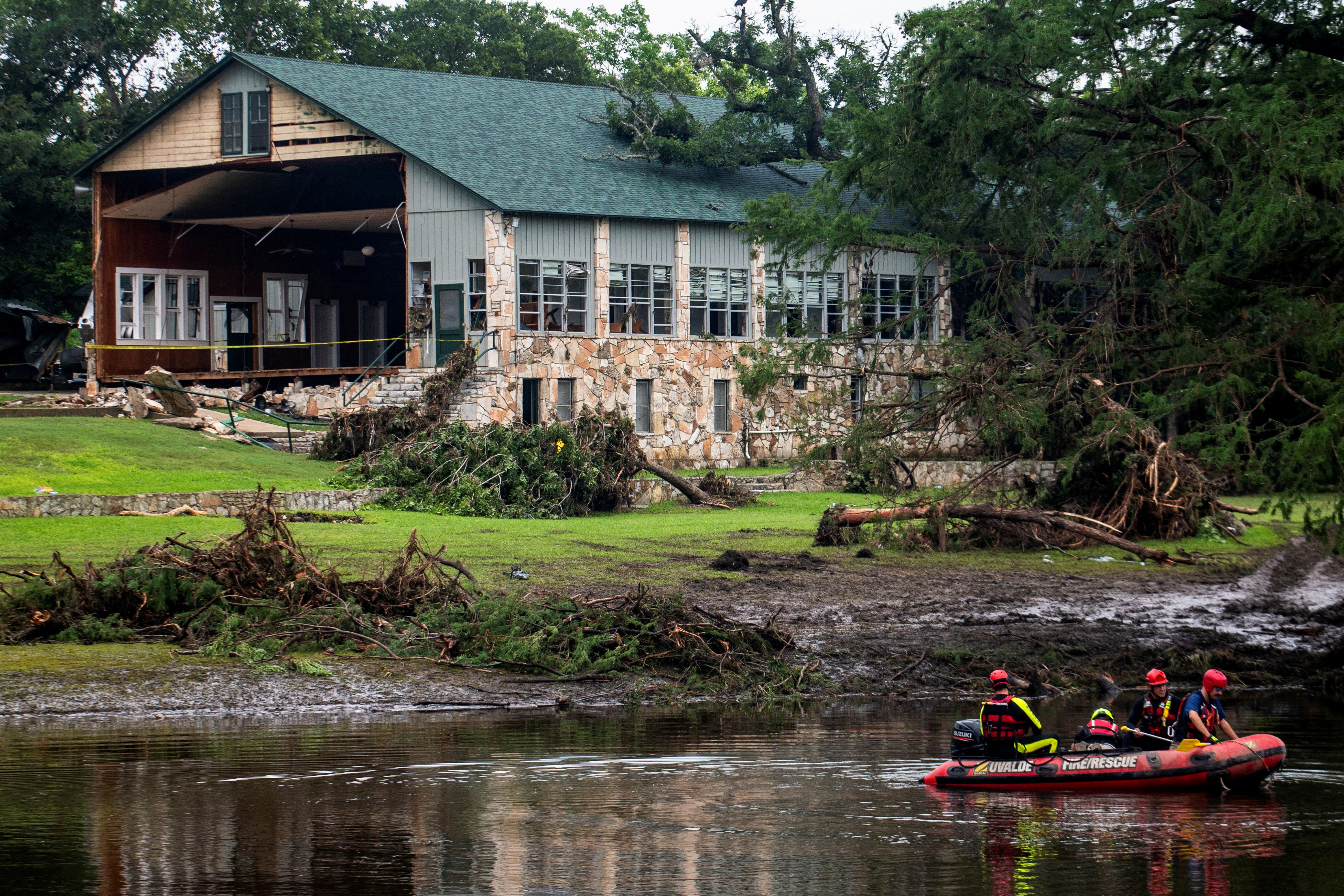 Rescue team in a red inflatable boat on water near a stone and wood building with severe damage and fallen trees, surrounded by muddy ground and debris after a storm.