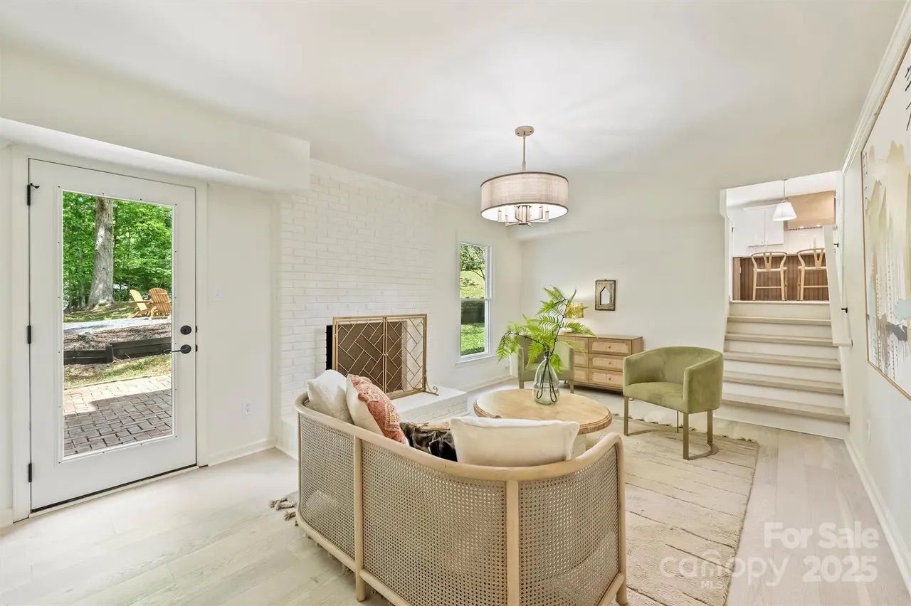 Bright living room with white walls and light wood floors, featuring a wicker-back curved sofa with cushions, a green armchair, round wooden coffee table with a plant, and a white brick fireplace.