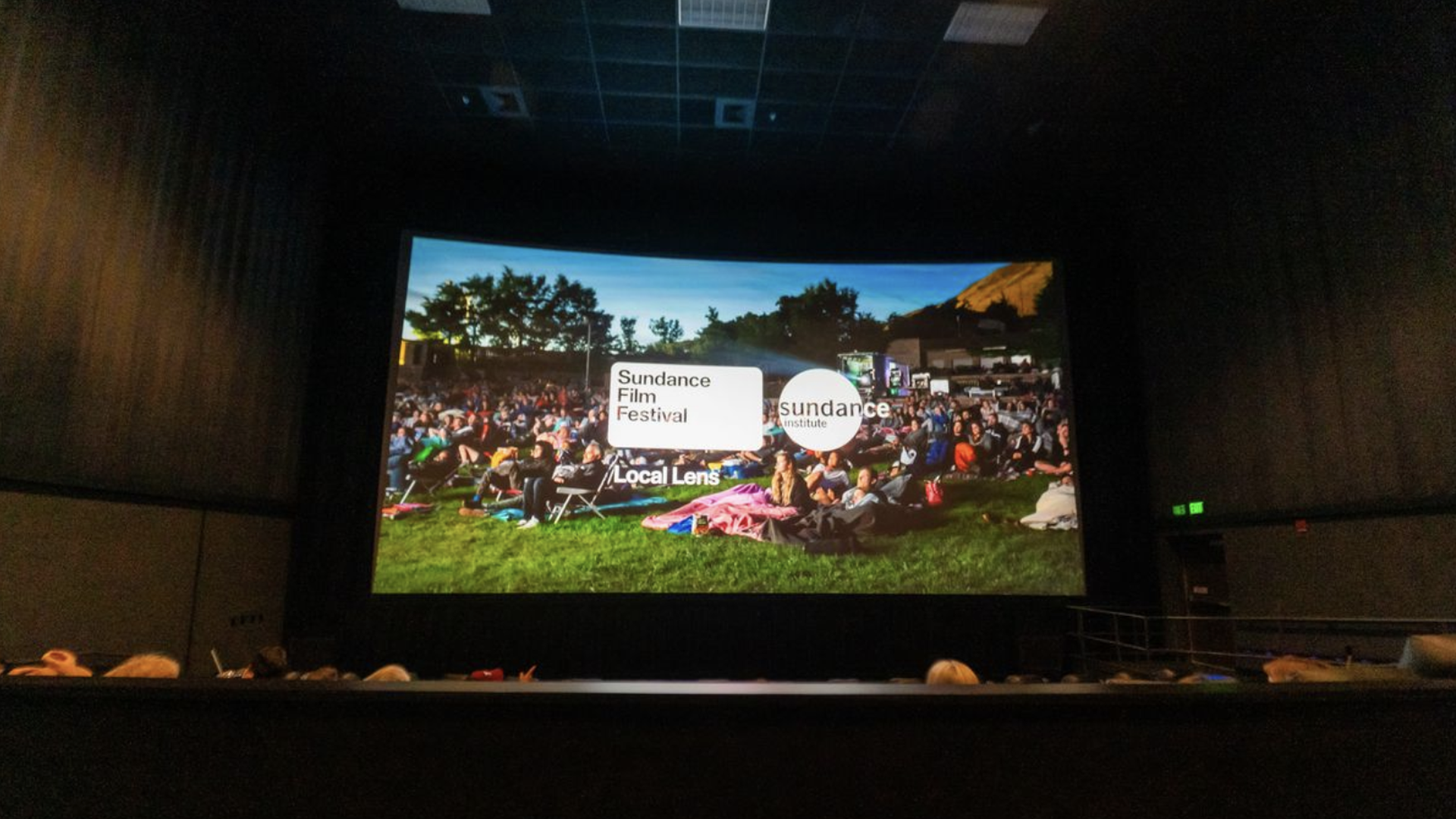 In a dark theater, a large screen shows an outdoor Sundance Film Festival crowd gathered in a park, with white text blocks reading "Sundance Film Festival" and "Local Lens".
