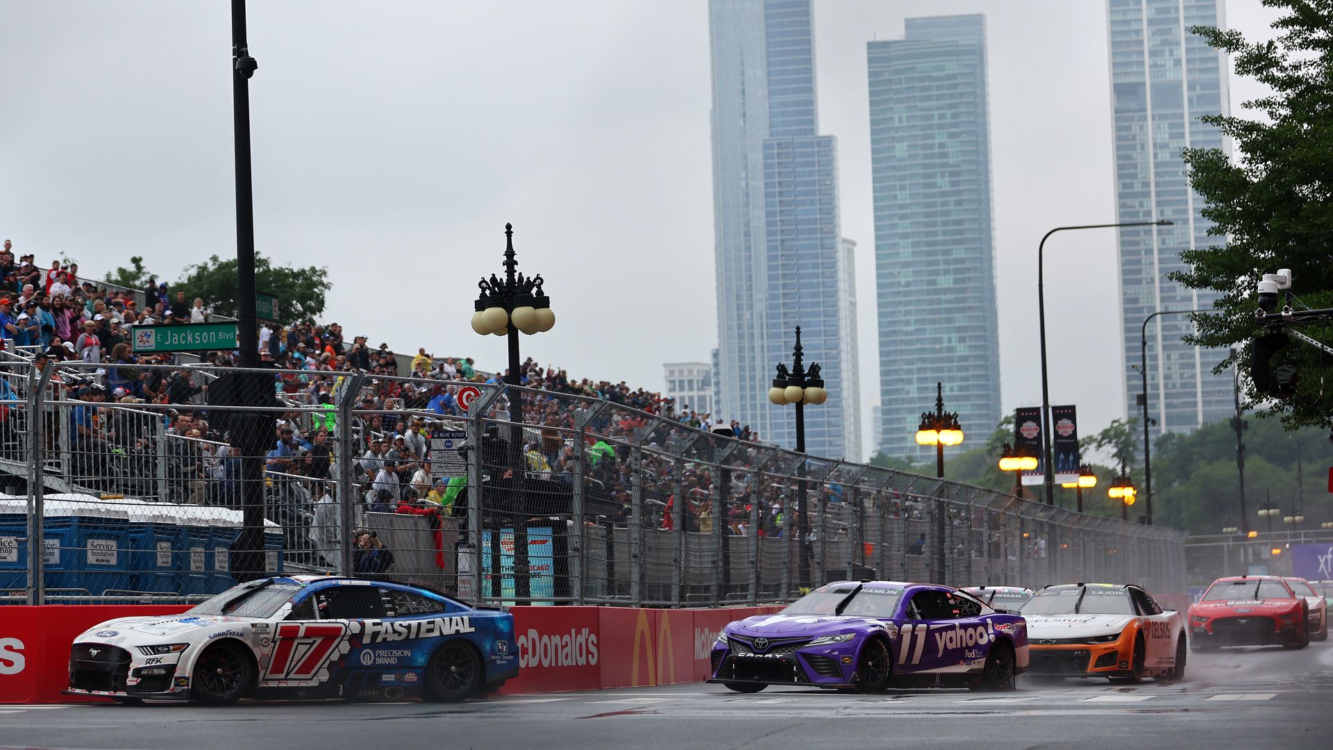 Race cars whip around a turn near stands of fans and the Chicago skyline in the background.