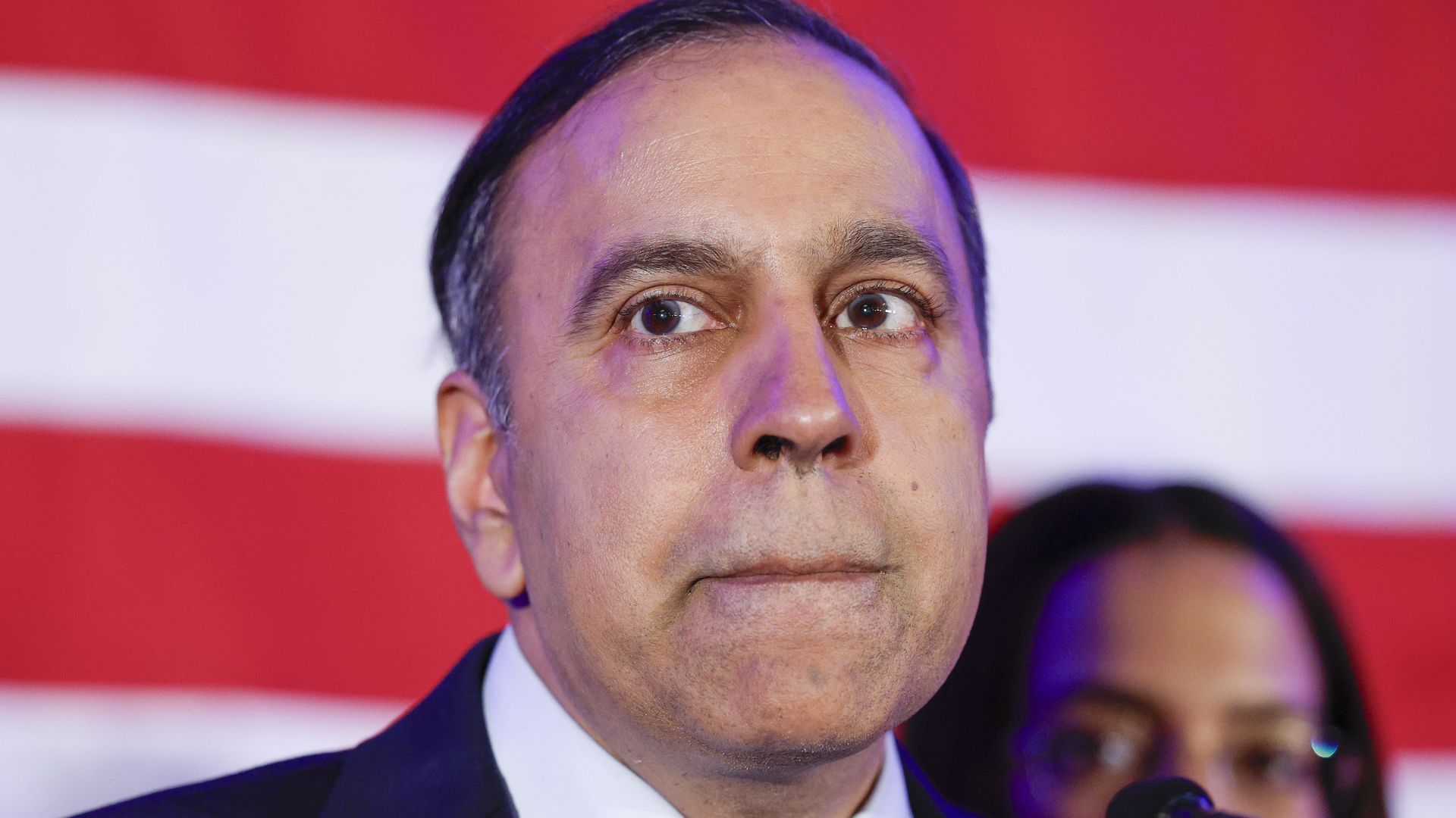 Close-up of a man in a dark suit and white shirt, looking to the side while speaking into a microphone; purple lighting on his face, with a red and white striped backdrop.