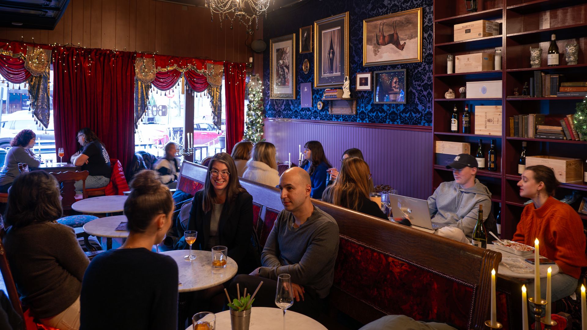 Cozy cafe scene with red velvet curtains, a chandelier, and blue damask wallpaper. People chat at round marble tables; a small Christmas tree glows near framed art and a bookshelf with bottles.