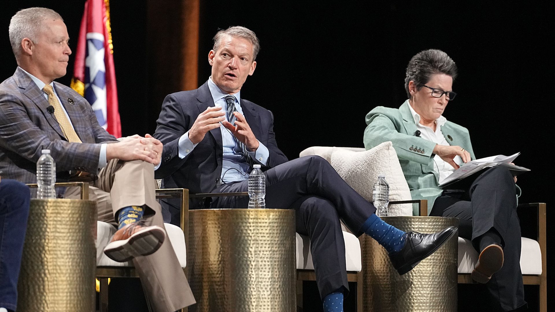 Three panelists on stage: left gray-haired man in plaid suit, center man in navy suit gesturing, right woman in green blazer reading papers; flag in background.