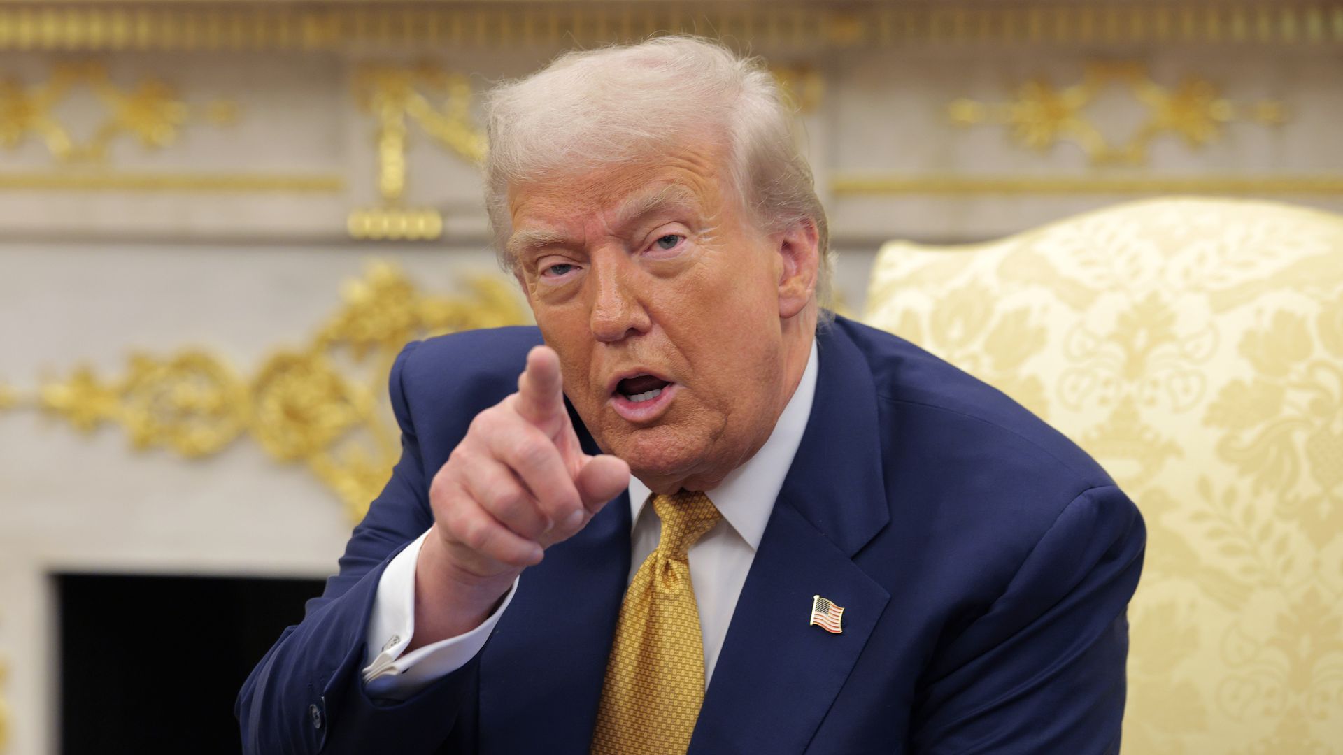 President Trump in a navy suit and yellow tie pointing forward, sitting on a beige patterned chair with gold decorations in the background.