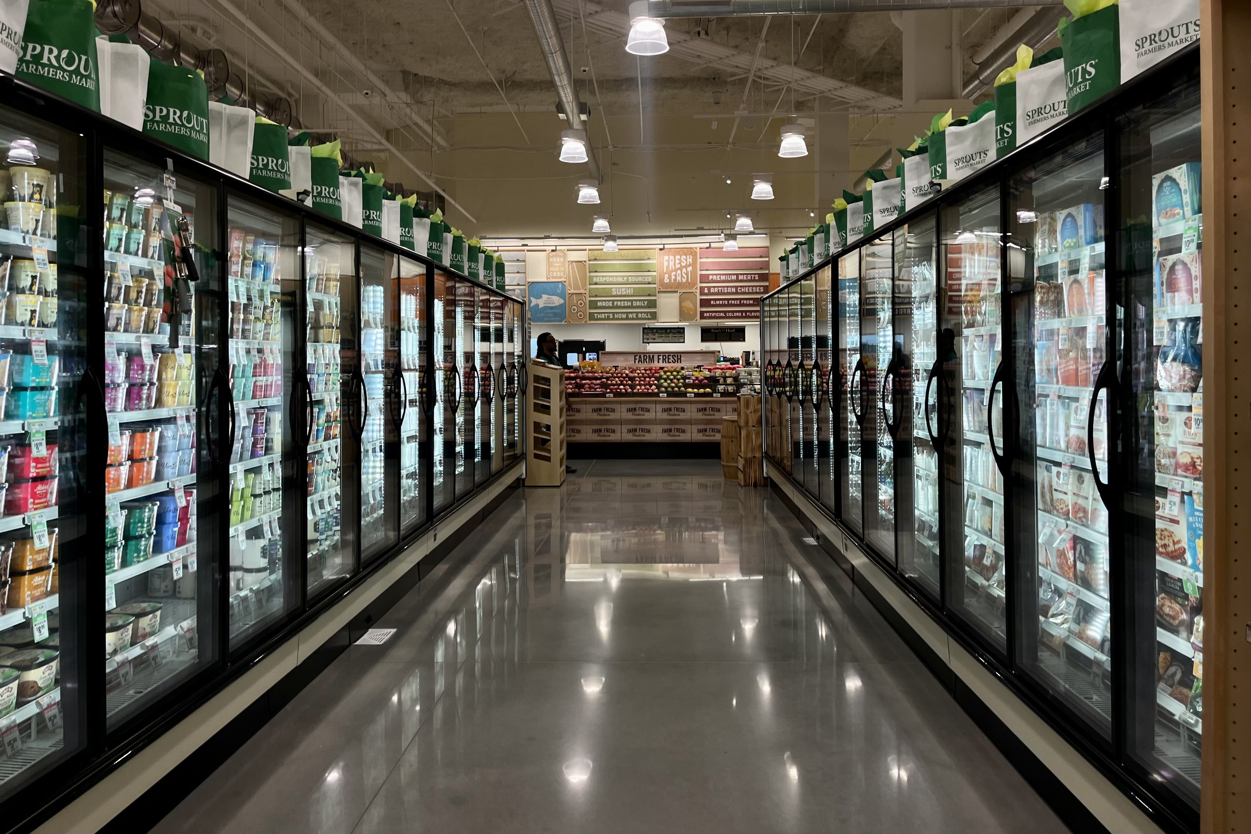 A grocery store aisle with glass-door freezers on both sides filled with various colorful frozen foods, and green-and-white Sprouts Farmers Market bags on top.