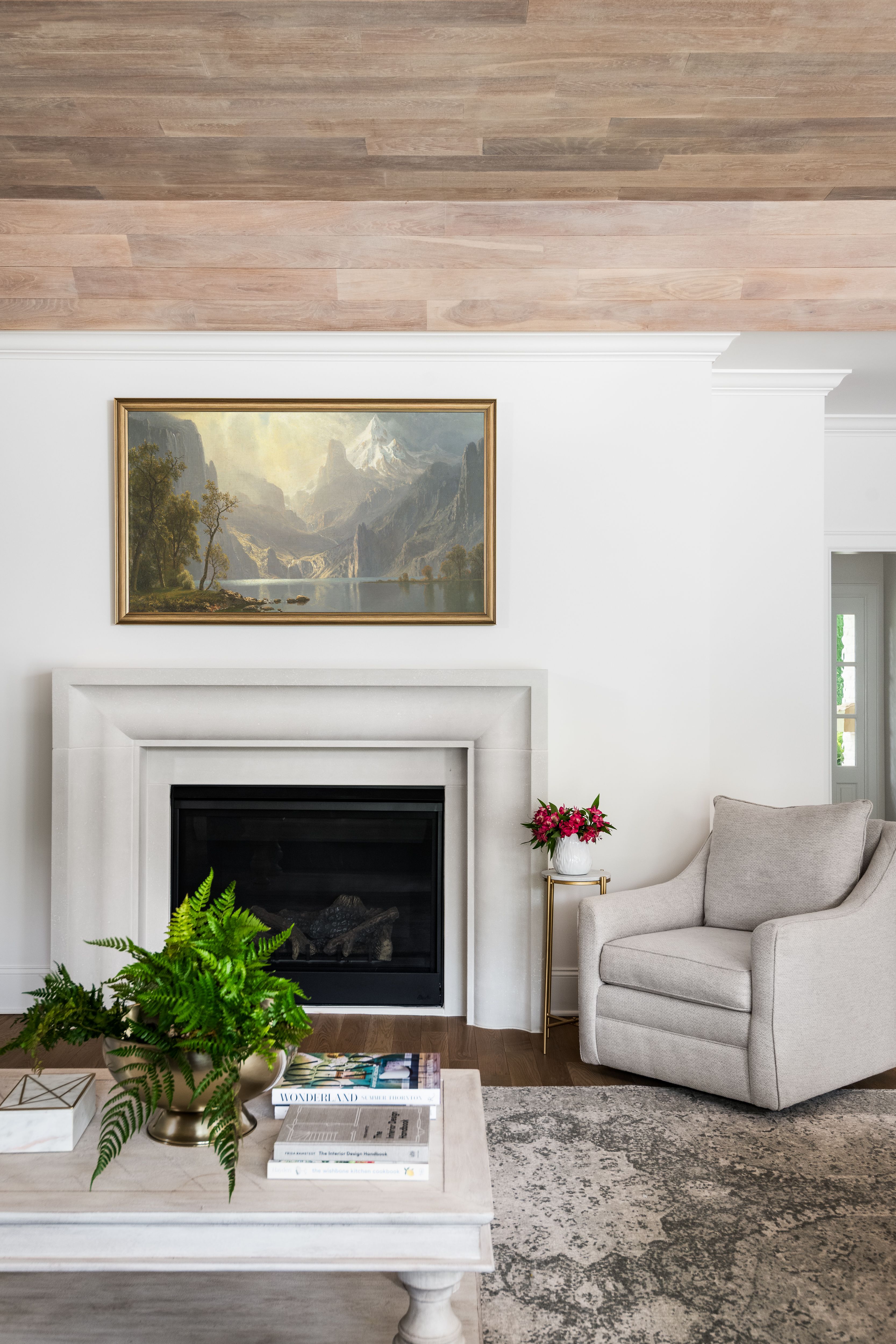 Cozy living room with a white fireplace, mountain landscape painting above, beige armchair, gold side table with pink flowers, rustic wood ceiling, light coffee table with green fern plant and books.