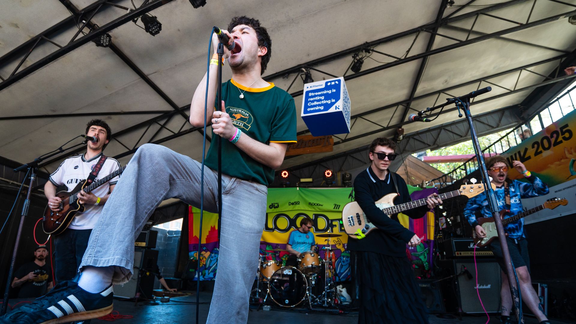 Band performing on stage under a tent, lead singer in green shirt and gray pants passionately singing into microphone, other members playing guitar and drums around colorful backdrop.