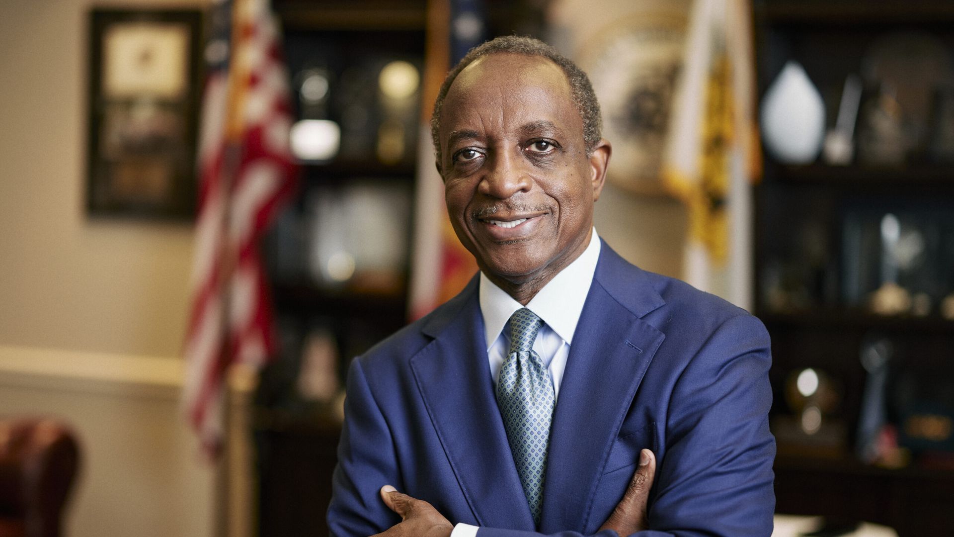 Smiling man in a blue suit and patterned tie standing with arms crossed in an office with American flags and shelves of awards and books in the background.