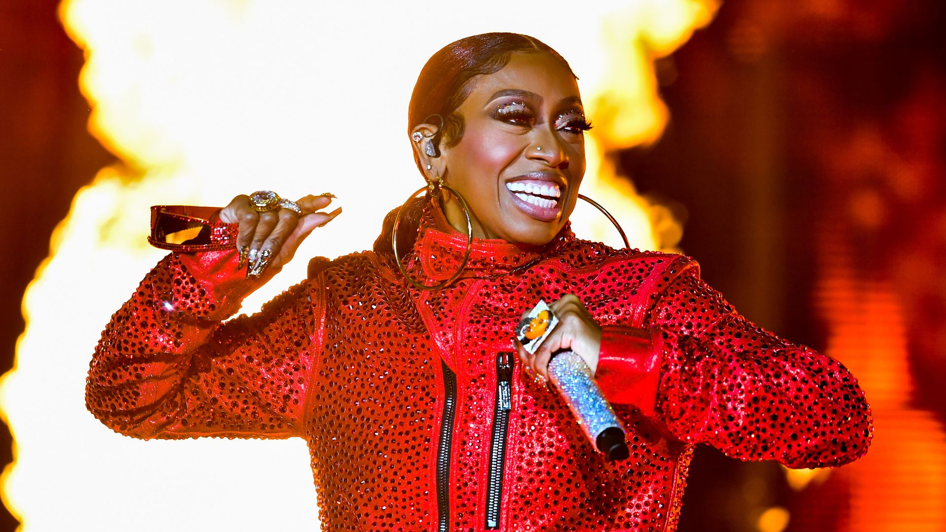 A woman onstage in a red outfit smiling while holding a mic with fire in the background.