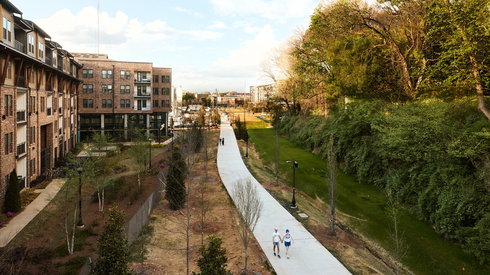 An aerial view of brick apartment buildings on the left, a white curved path through a park in the center, two runners in the foreground, and dense trees on the right under a blue sky.