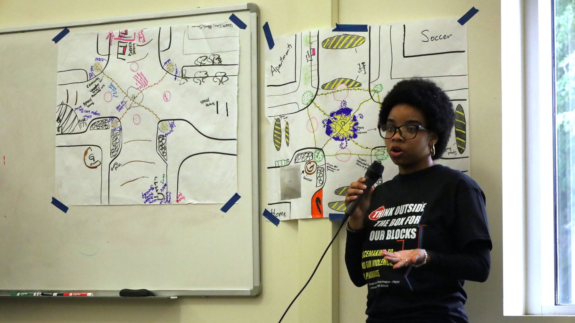 A young woman speaking into a microphone, standing in front of two pieces of paper with drawings of an intersection on them taped to the wall.