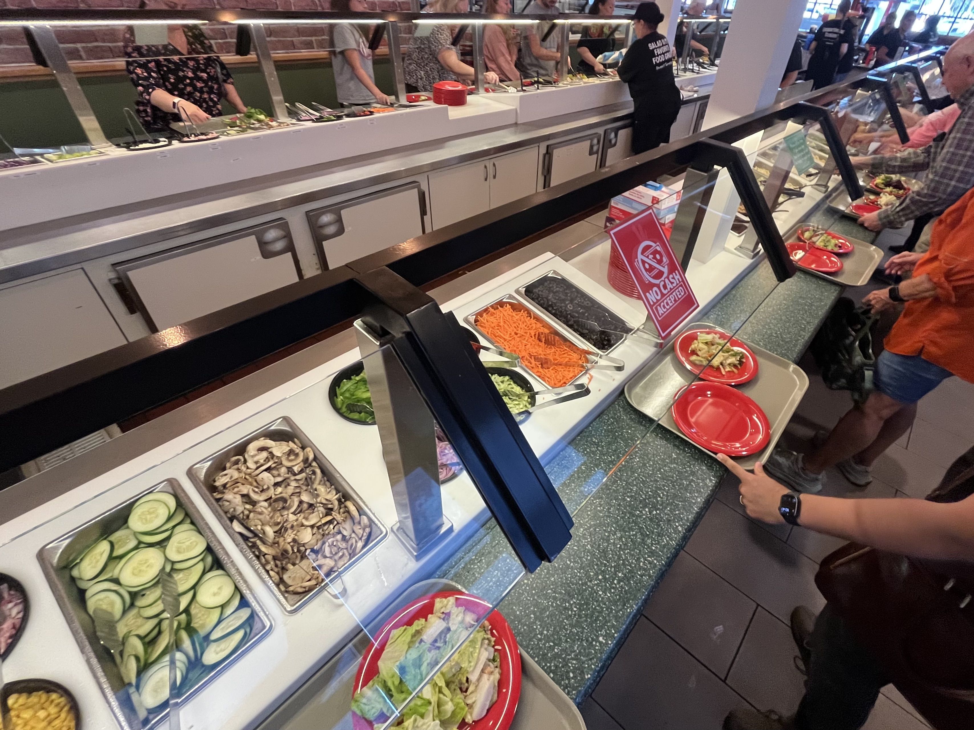Long cafeteria line with a serving counter and sneeze guard; people select salads from trays of cucumbers, mushrooms, carrots, and greens. Red plates and a "No cash accepted" sign visible.