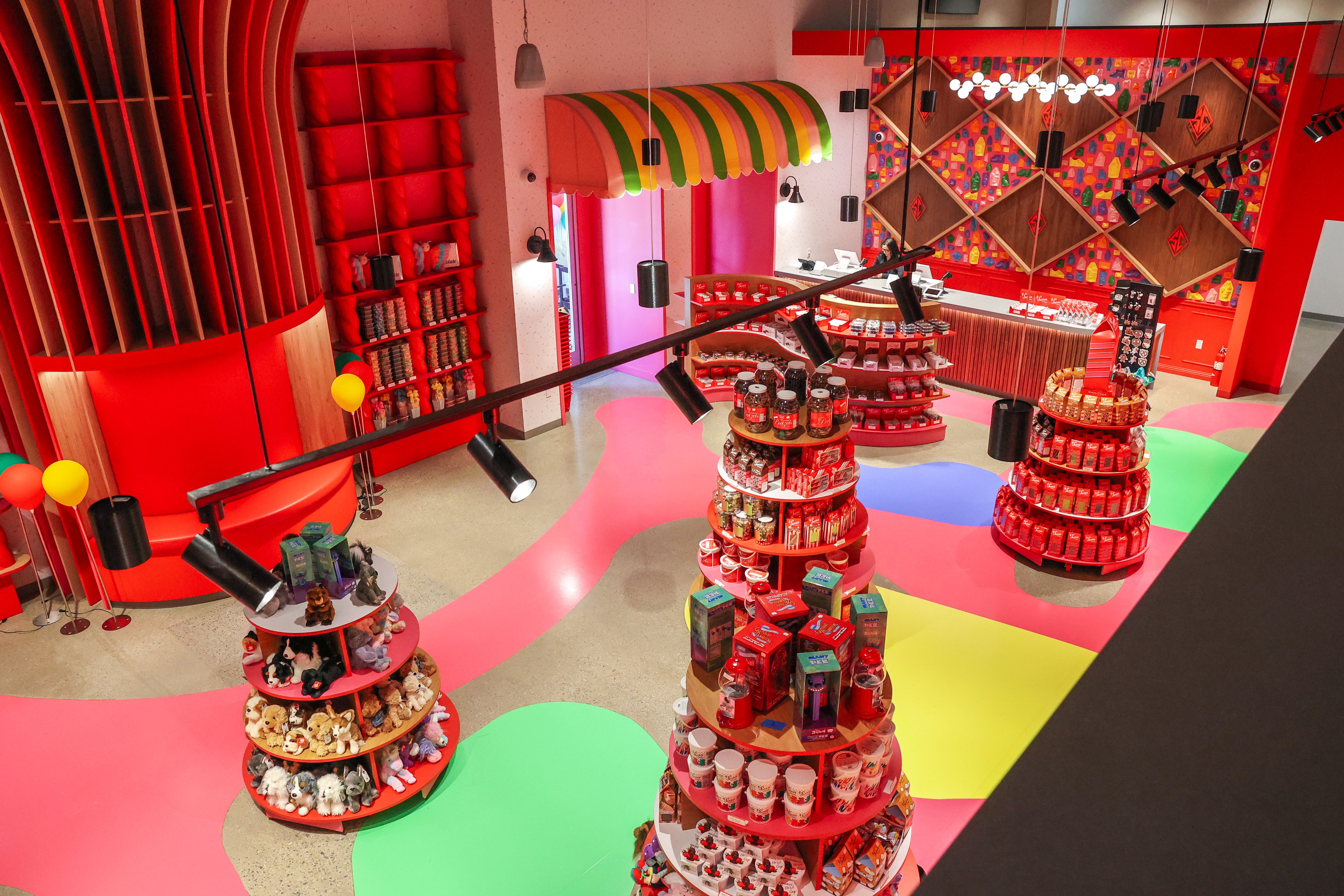 Colorful candy shop interior with red walls and curved shelves. Central round displays hold jars and treats, pink and green floor paths, rainbow striped canopy, and a geometric mural on the back wall.