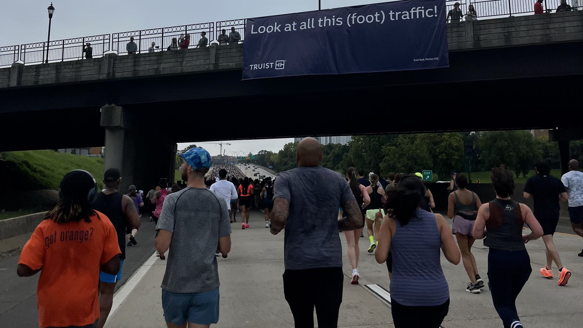 Runners fill a city street under a bridge, where a large blue banner says "Look at all this (foot) traffic!" Spectators watch from the overpass as the race continues.