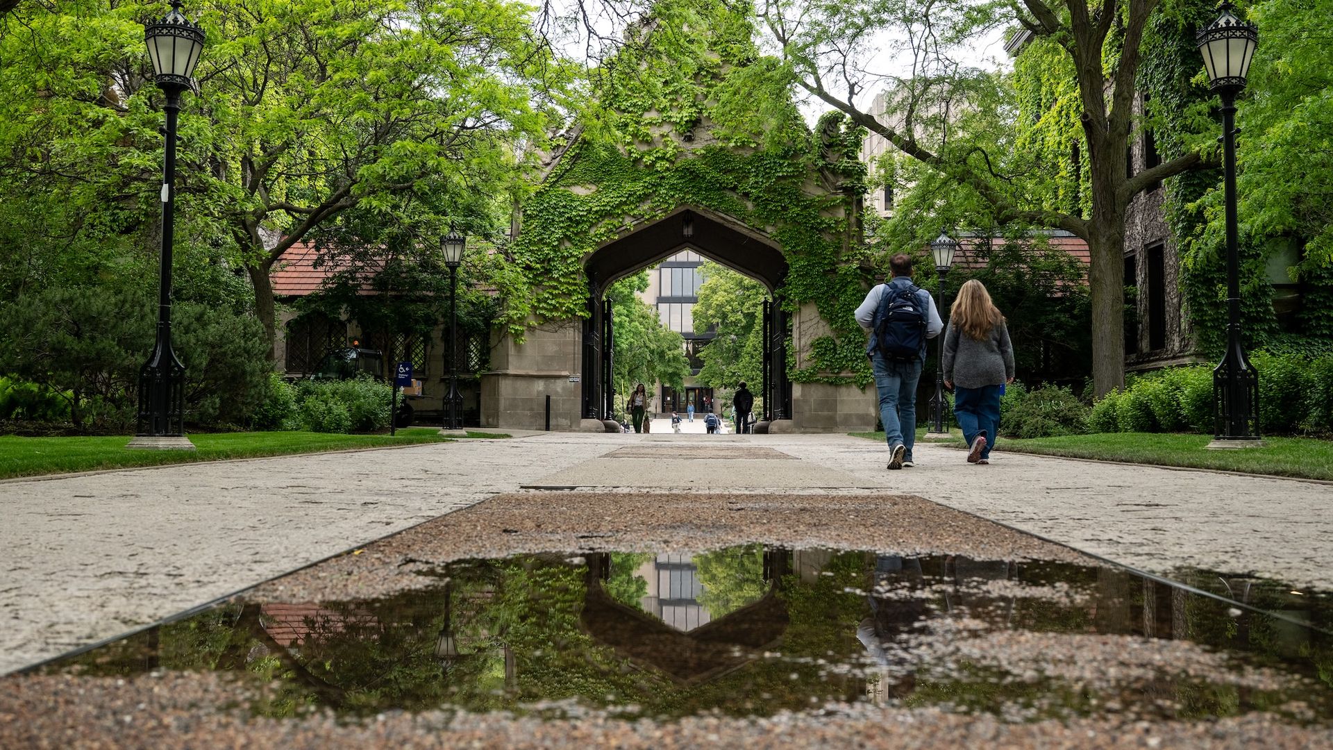 Tree-lined University of Chicago campus path toward an ivy-covered stone archway; ornate black street lamps flank the walkway; two people with backpacks walk toward the arch, reflected in a puddle on the path.
