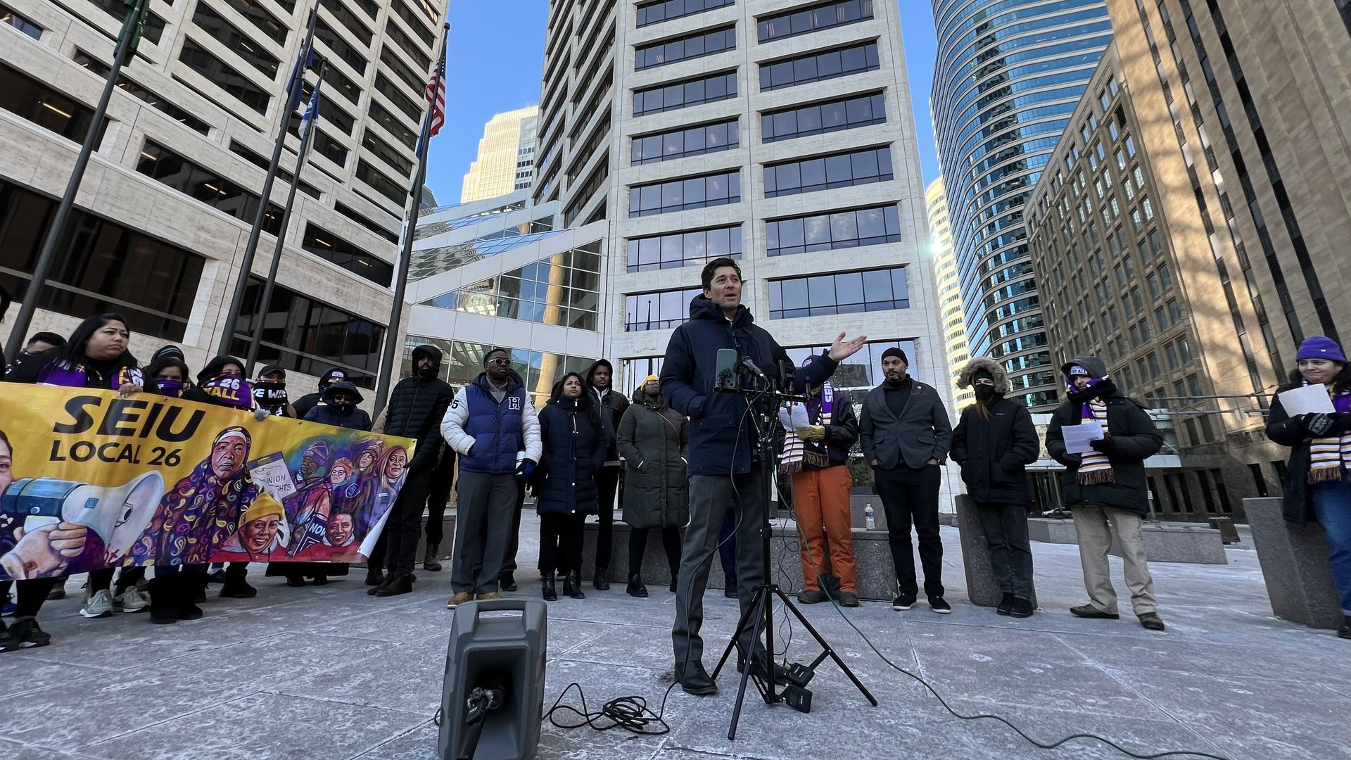 A man with light skin in a black jacket speaks at a microphone stand at a press conference in an urban downtown in front of a white skyscraper