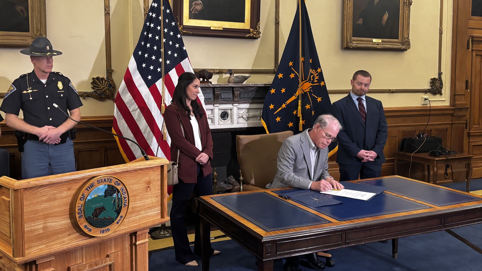 A formal signing ceremony in a wood-paneled room. A man signs documents at a desk while a woman and two men stand nearby; US and Indiana flags and portraits adorn the background.