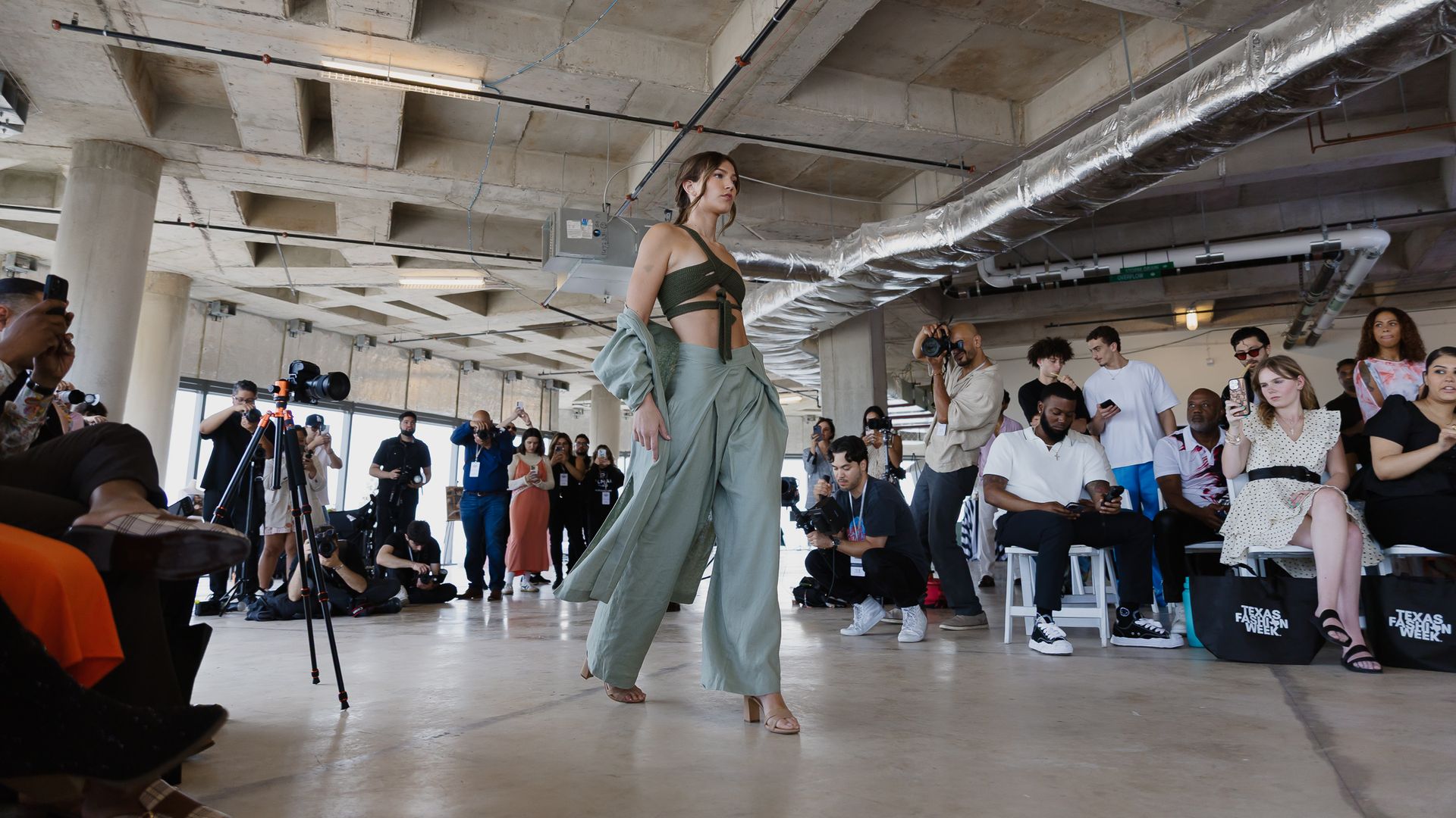 Model walks a fashion show in an industrial loft, wearing a green draped outfit; audience, photographers, and cameras line the runway, with exposed concrete ceiling and metallic ducts.