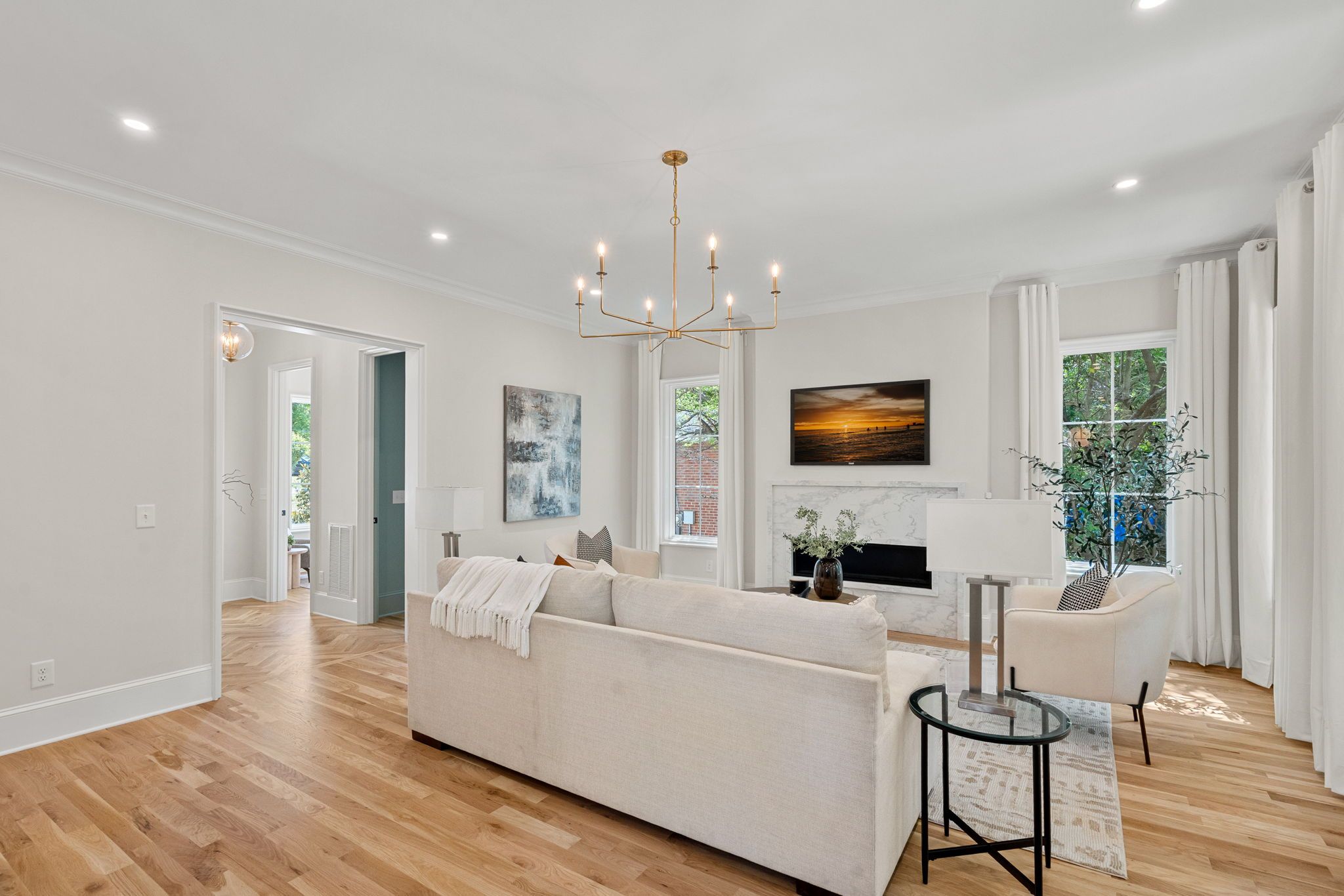 Bright living room with light wood floors, beige sofa, glass round coffee table, white curtains, and a gold multi-arm chandelier. TV above a marble fireplace, blue artwork, and plants by the windows.