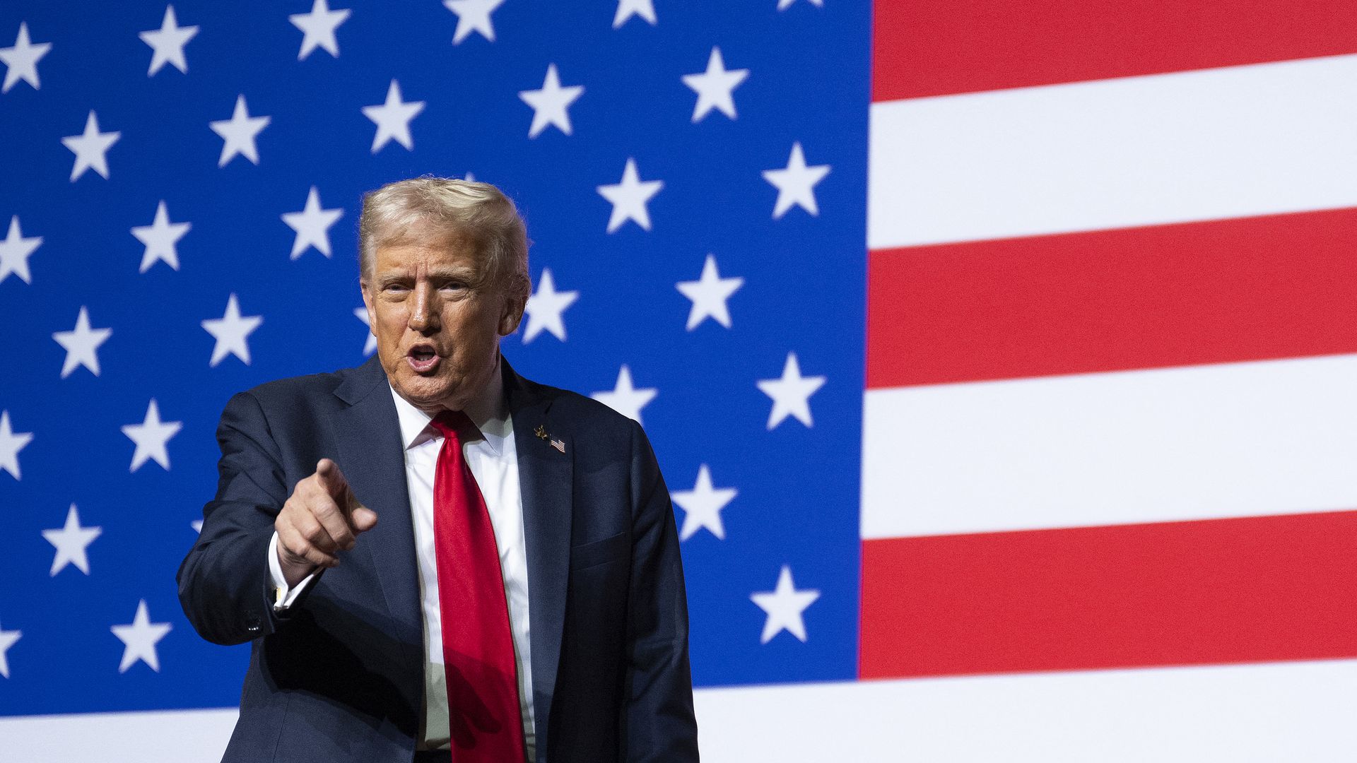 President Trump, wearing a navy jacket with a US flag, white shirt and red tie, points with his right hand in front of a US flag as he speaks.