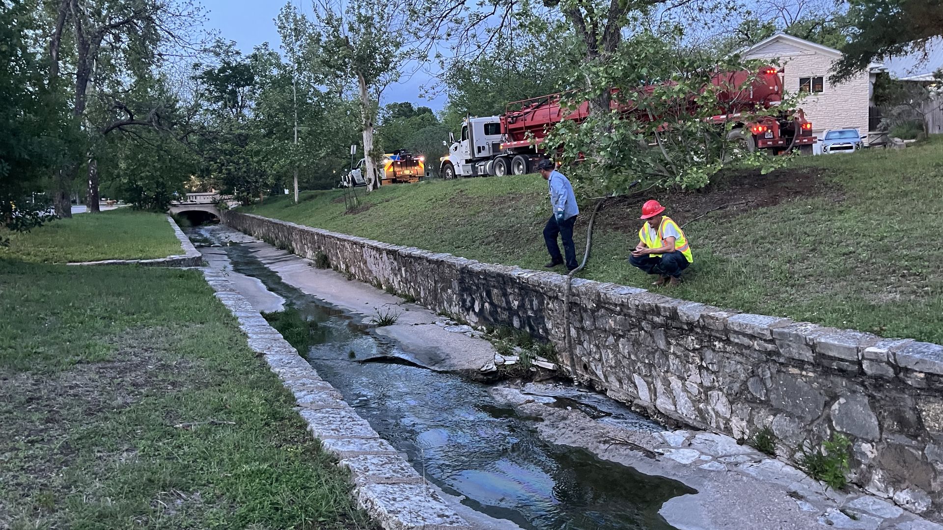 Two construction workers in safety vests and hard hats repair a concrete, stone-walled drainage canal along a grassy verge. A large red vacuum truck and a residential house are in the background.