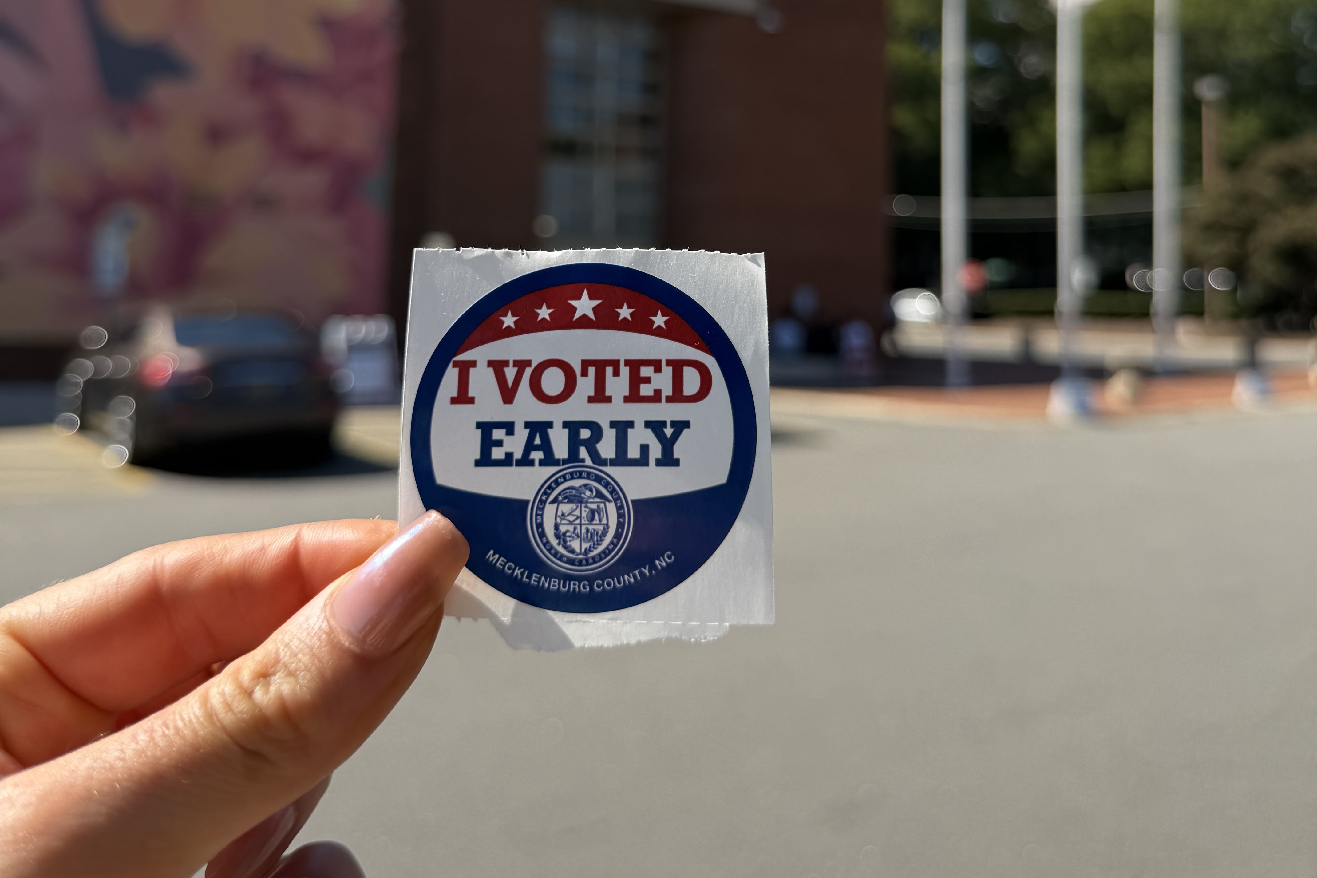 A close-up of a hand holding an I Voted Early sticker with red and blue text and a circular seal; the background shows a blurred parking lot and brick building.