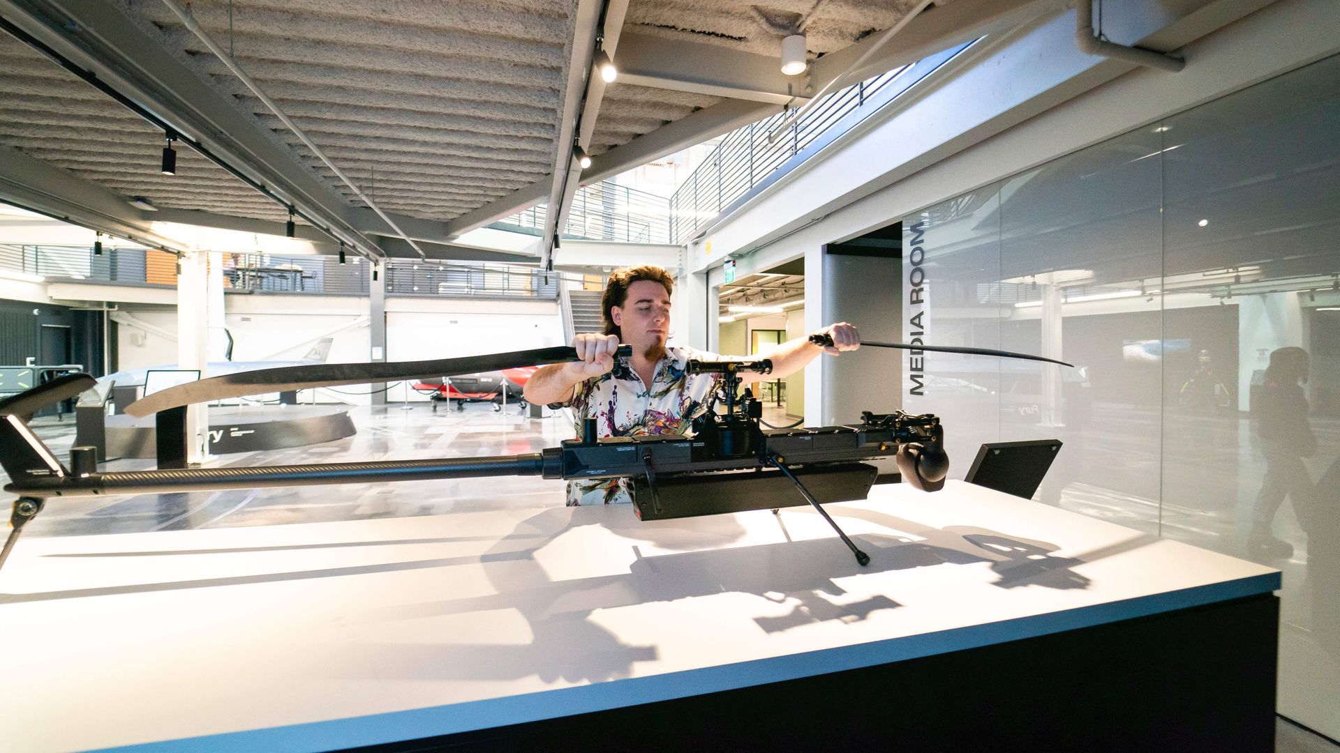 A man, Palmer Luckey, affixes black rotor blades onto a drone in a facility. The walls and doors are shiny white, with a grey ceiling overhead.