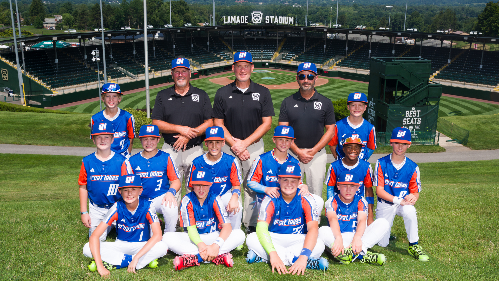 Young baseball players in blue jerseys and matching caps pose with three coaches on the grassy field in front of Lamade Stadium, with the diamond and empty stands behind.