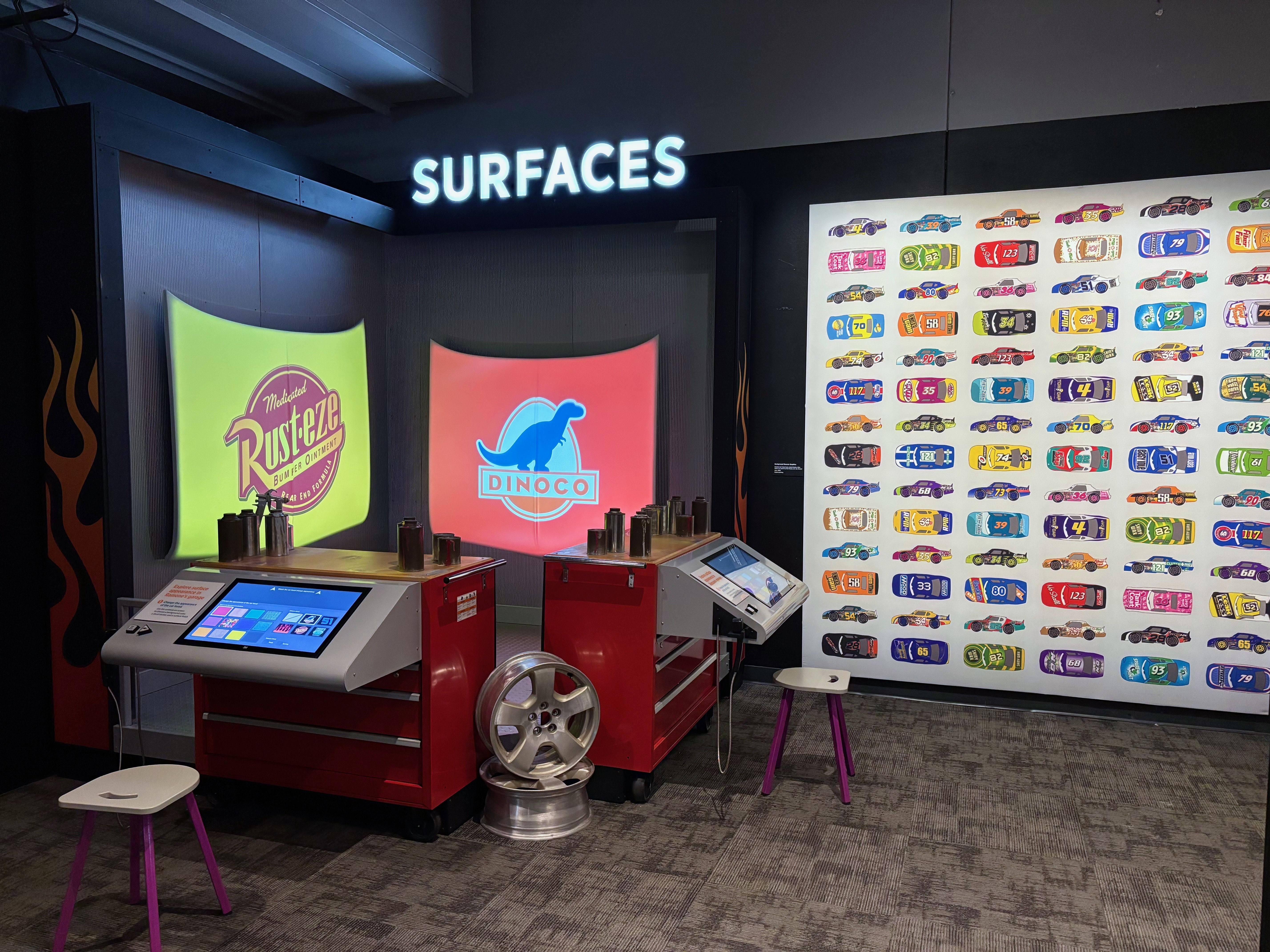 Interactive exhibit titled SURFACES with Rust-eze and Dinoco logos illuminated, red tool carts with screens and wheels, and a wall displaying colorful race car designs.