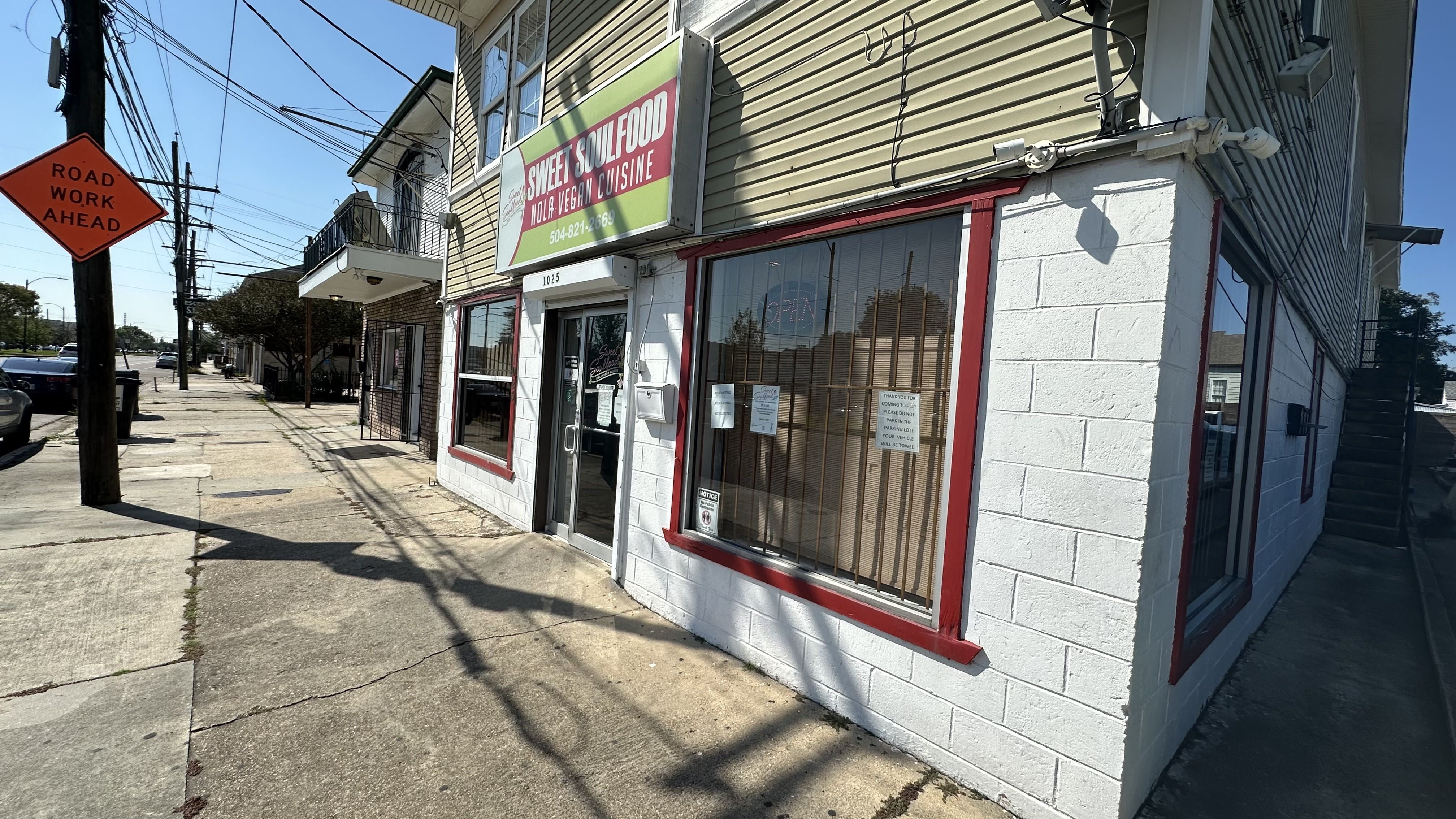 Street view of a beige and white building with red window frames housing Sweet Soulfood, a vegan cuisine restaurant, under a green sign. Orange "Road Work Ahead" sign on sidewalk.