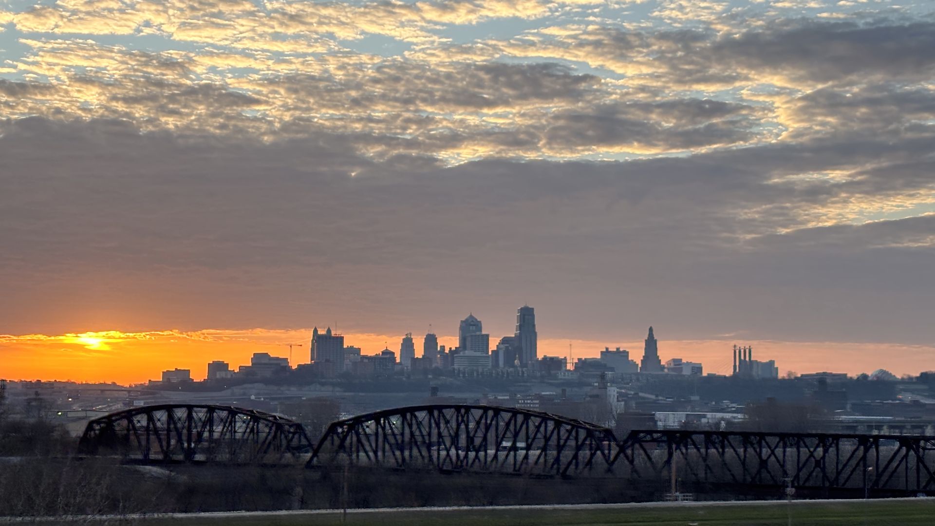 Sunrise over a city skyline with orange horizon and silhouetted skyscrapers; dark lattice-truss bridges in the foreground and a cloudy sky with golden highlights.