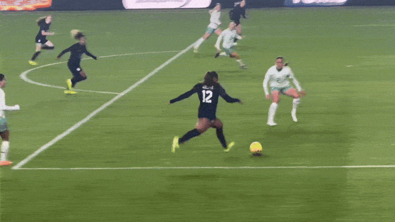 Soccer player wearing black jersey number 12 kicks a yellow ball toward the goal while players in white jerseys defend on a green field during a match.