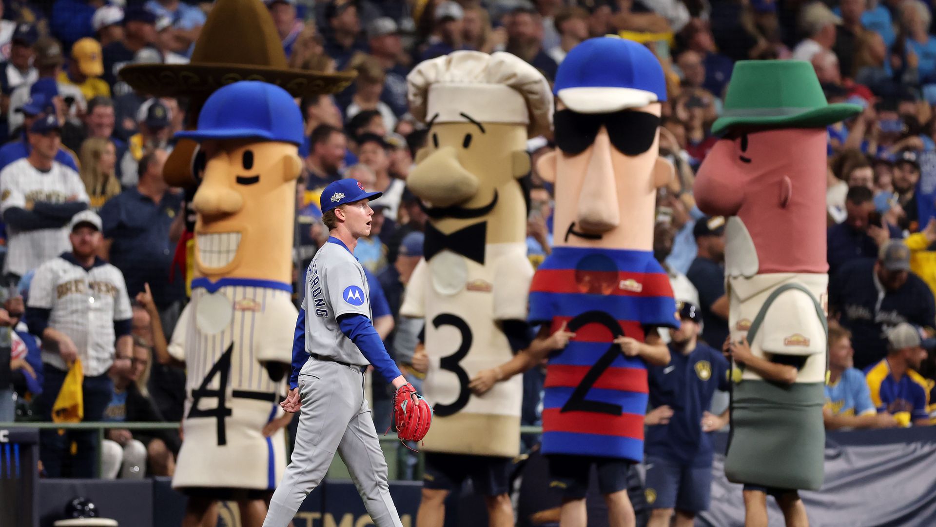Photo of a baseball player in uniform walking past four sausage-shaped mascots at a baseball stadium. 