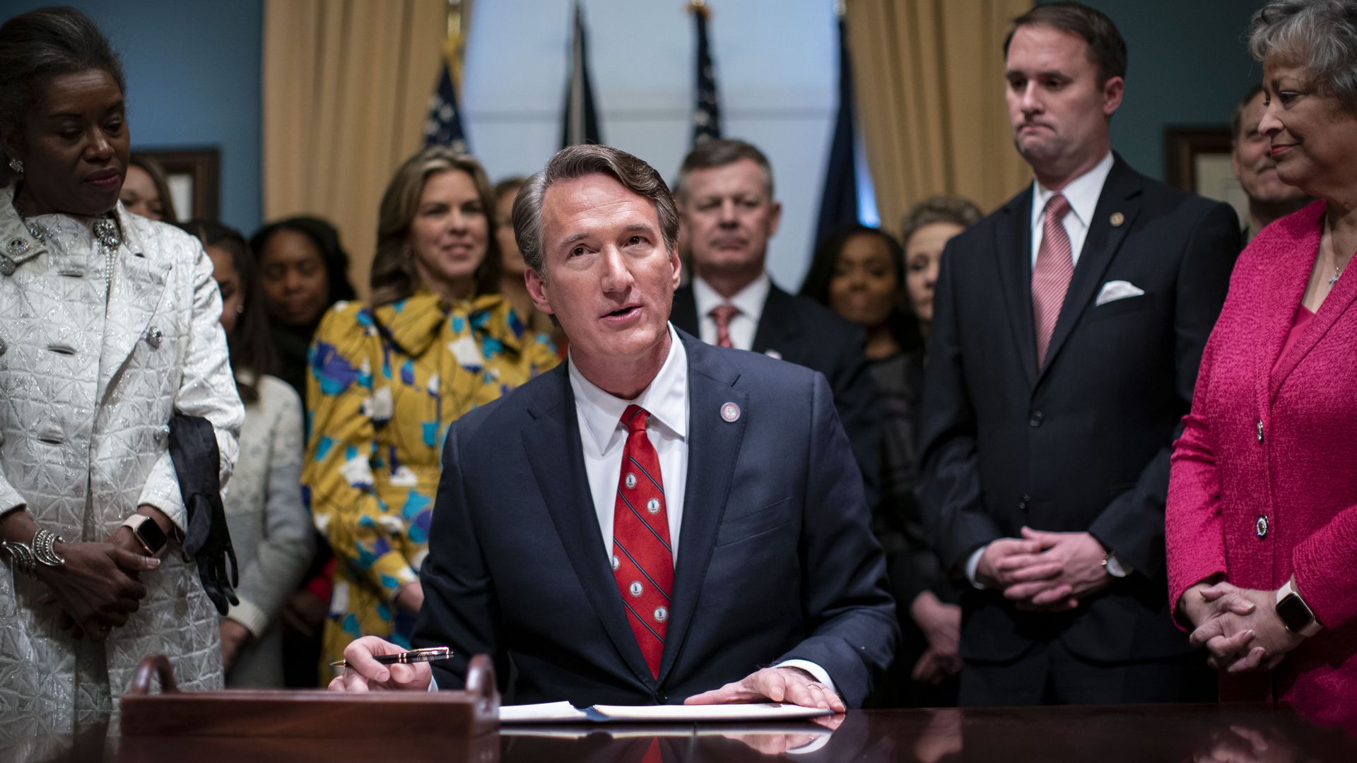 Glenn Youngkin, center, is seated at desk signing executive orders, surrounded by cabinet.