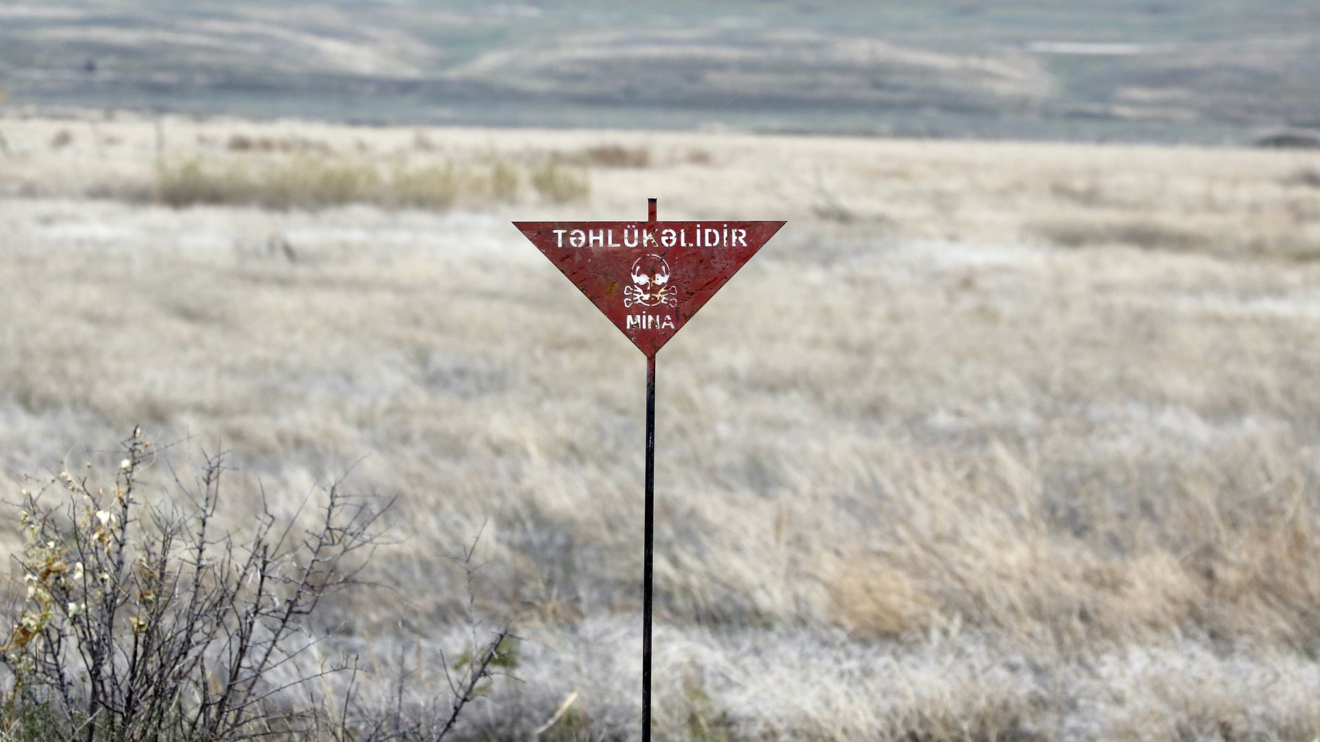 A red inverted triangle warning sign with a skull and crossbones sits on a thin post in a dry, open field, with distant grassy hills and a pale blue sky.
