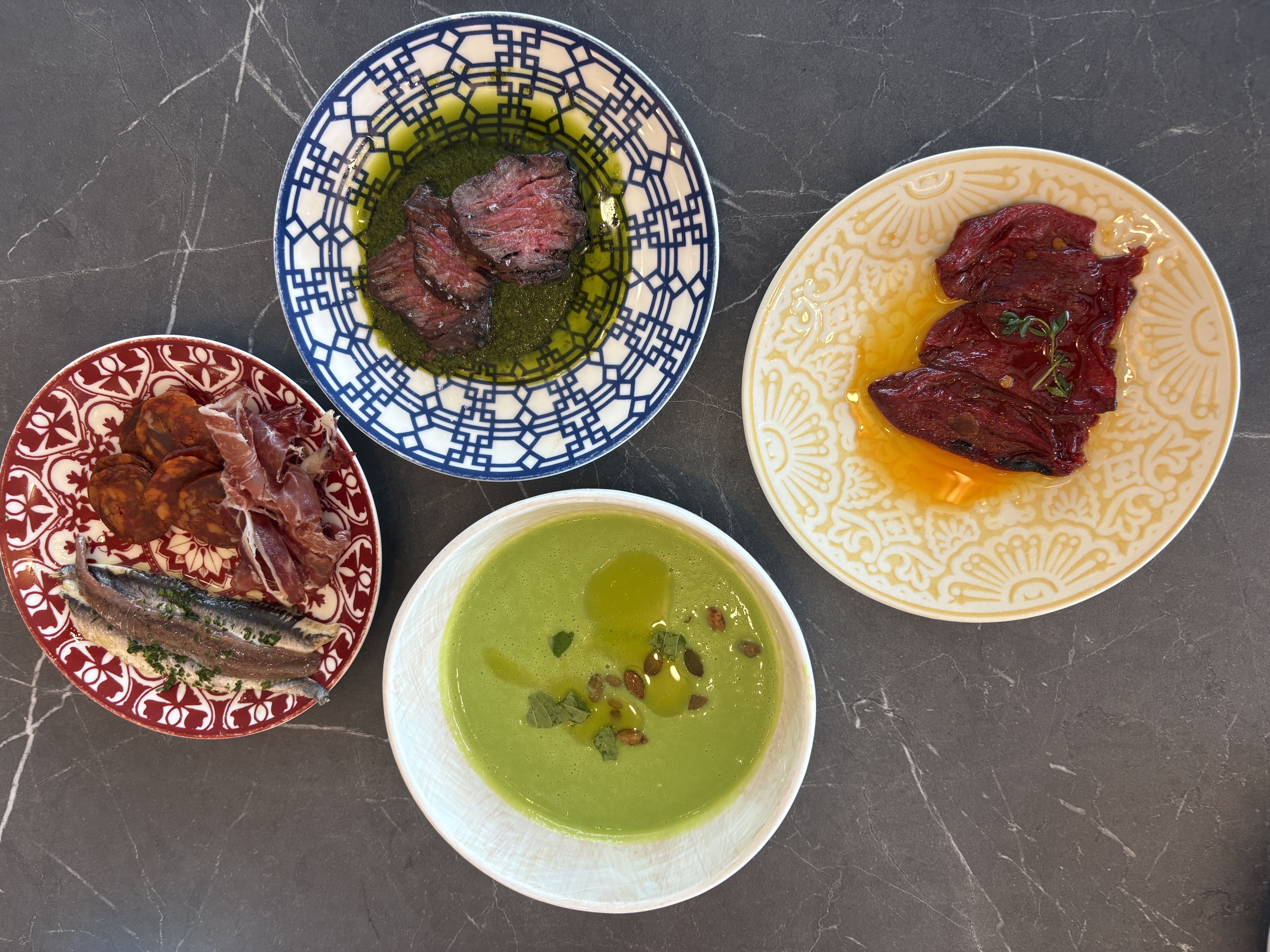 Top-down view of a dark marble table with several bowls of food: blue geometric bowl with sliced beef in green oil, yellow-patterned plate with red meat in yolk, red patterned plate of sliced meats, and a green soup.