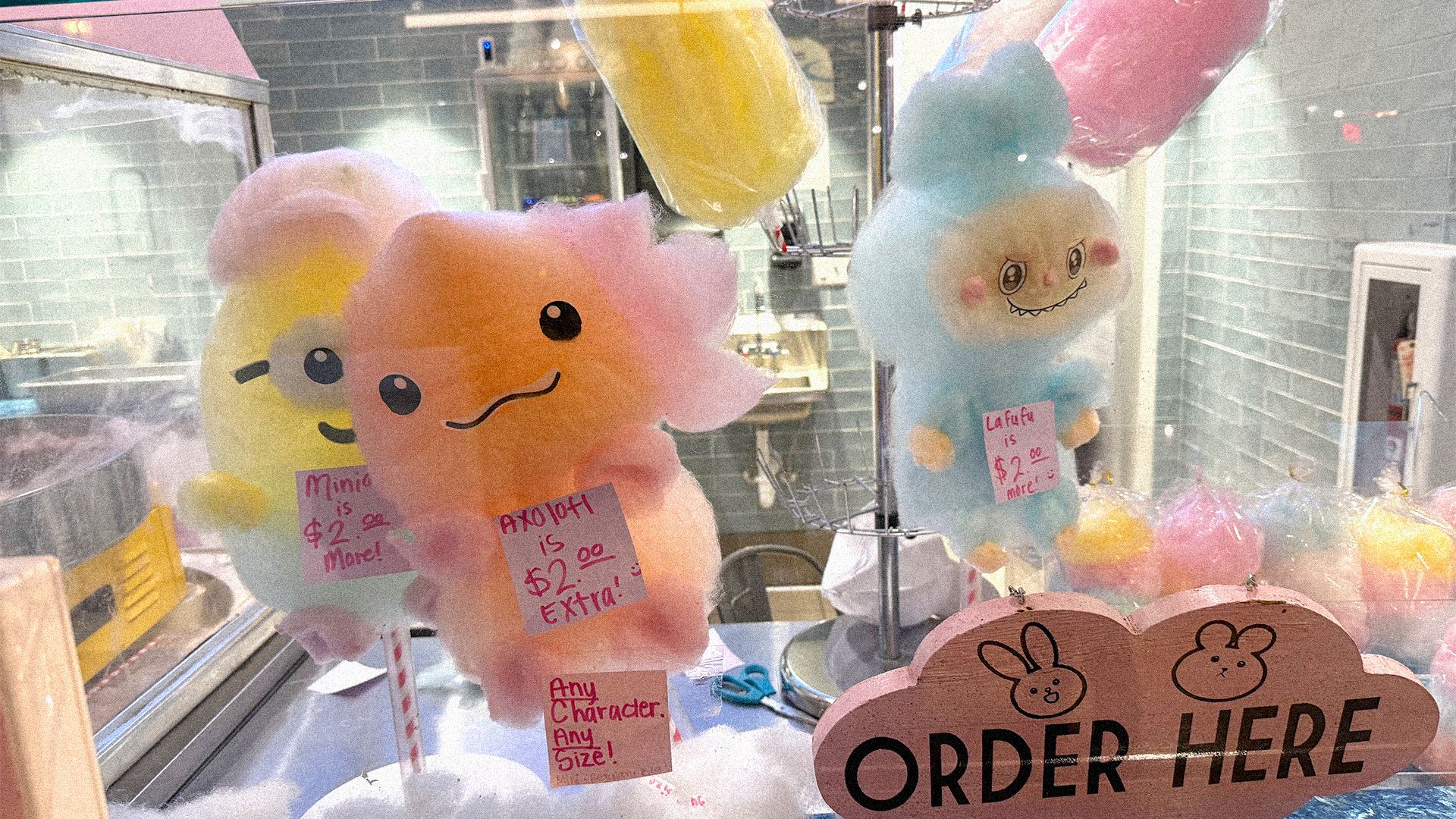 Colorful cotton candy shaped like cute characters with faces displayed behind a glass counter, featuring pink, blue, yellow, and orange hues and signs with prices and "ORDER HERE" wooden sign.