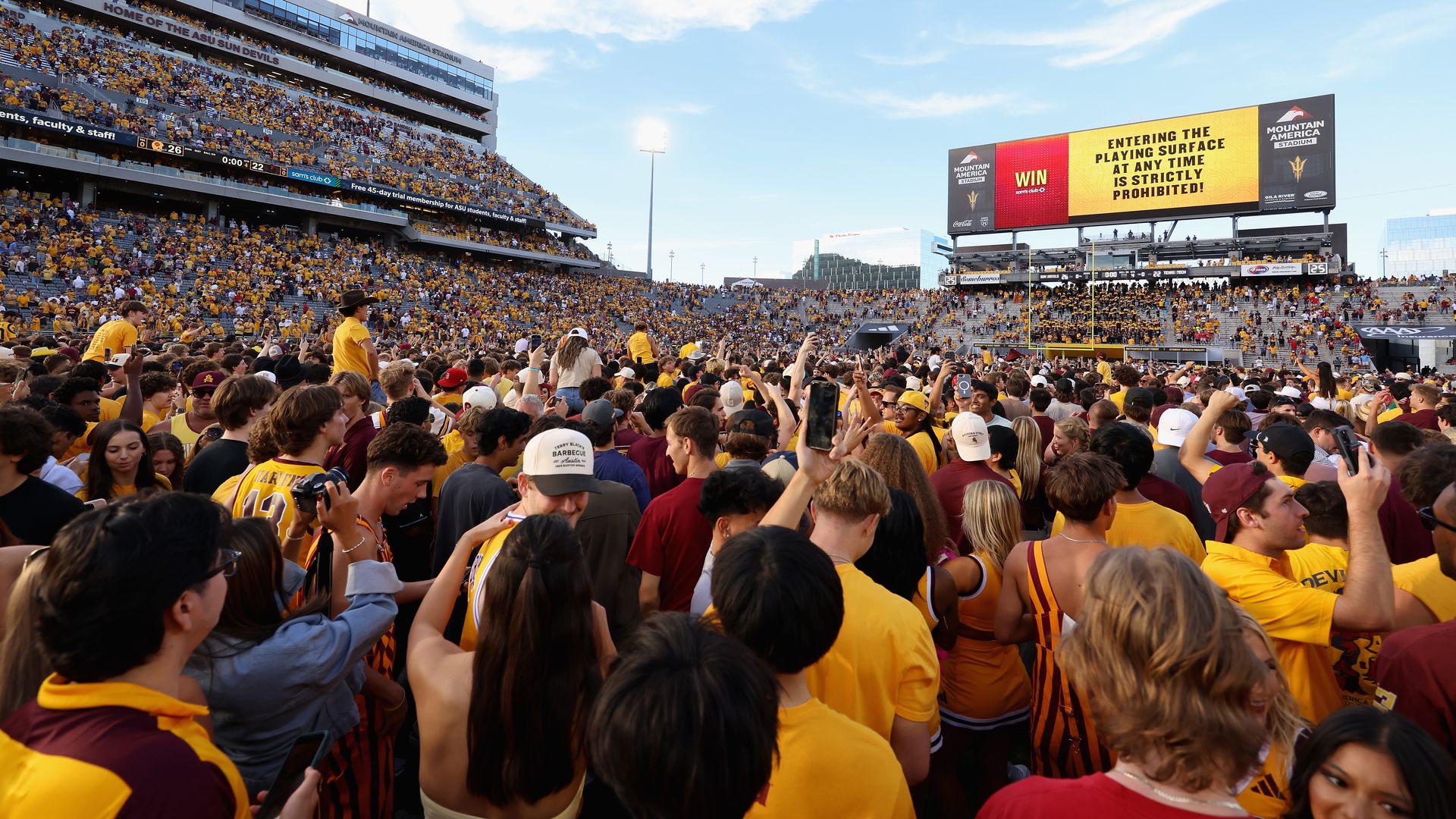 Large crowd of fans, mostly in yellow and maroon, celebrating on a football field at Mountain America Stadium under a partly cloudy sky with a scoreboard displaying rules and sponsor ads.