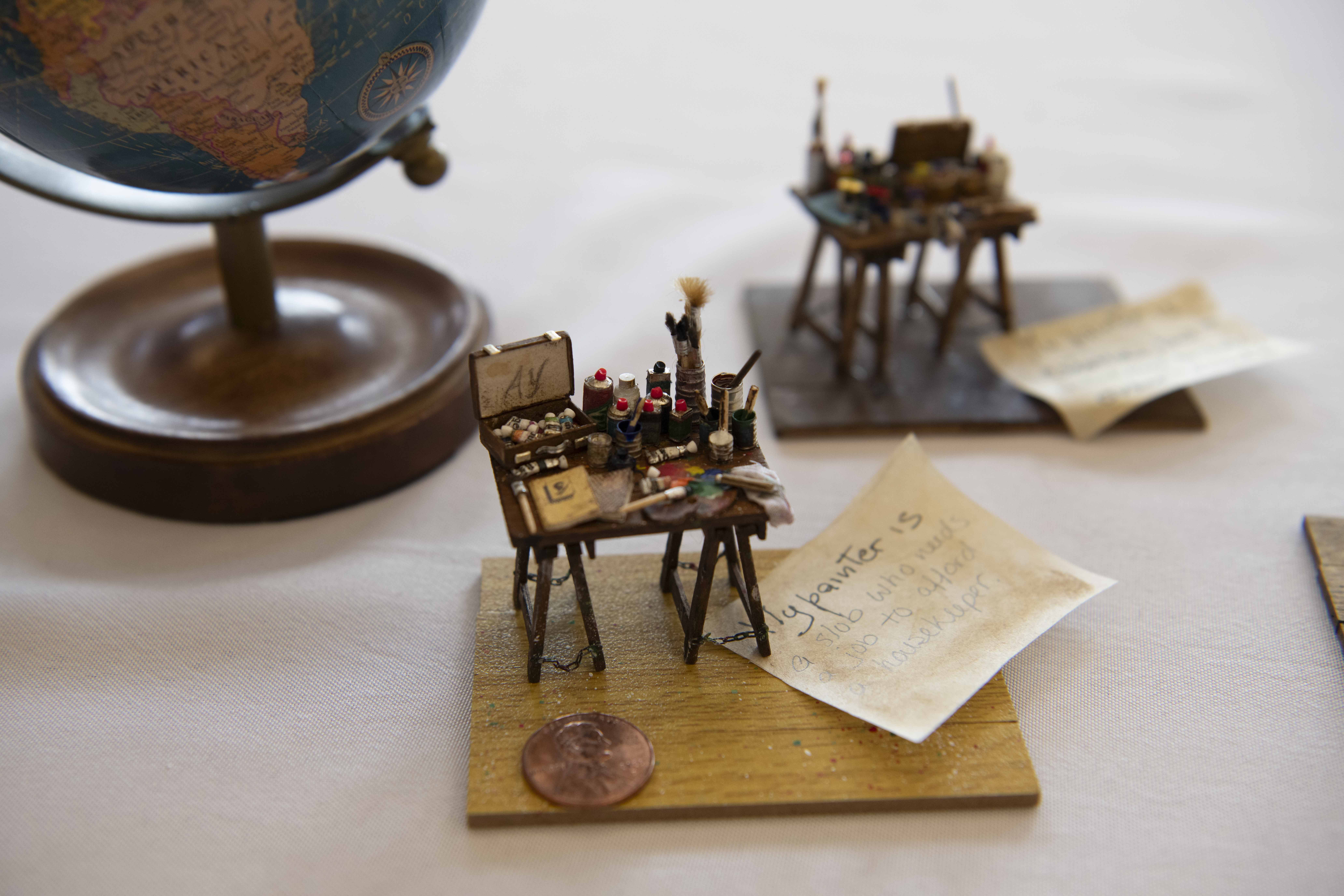 Globe on a wooden stand to the left, with tiny easels, paint tubes, and brushes on a light table. Handwritten notes and a penny lie nearby; a blurred backdrop shows more art supplies.