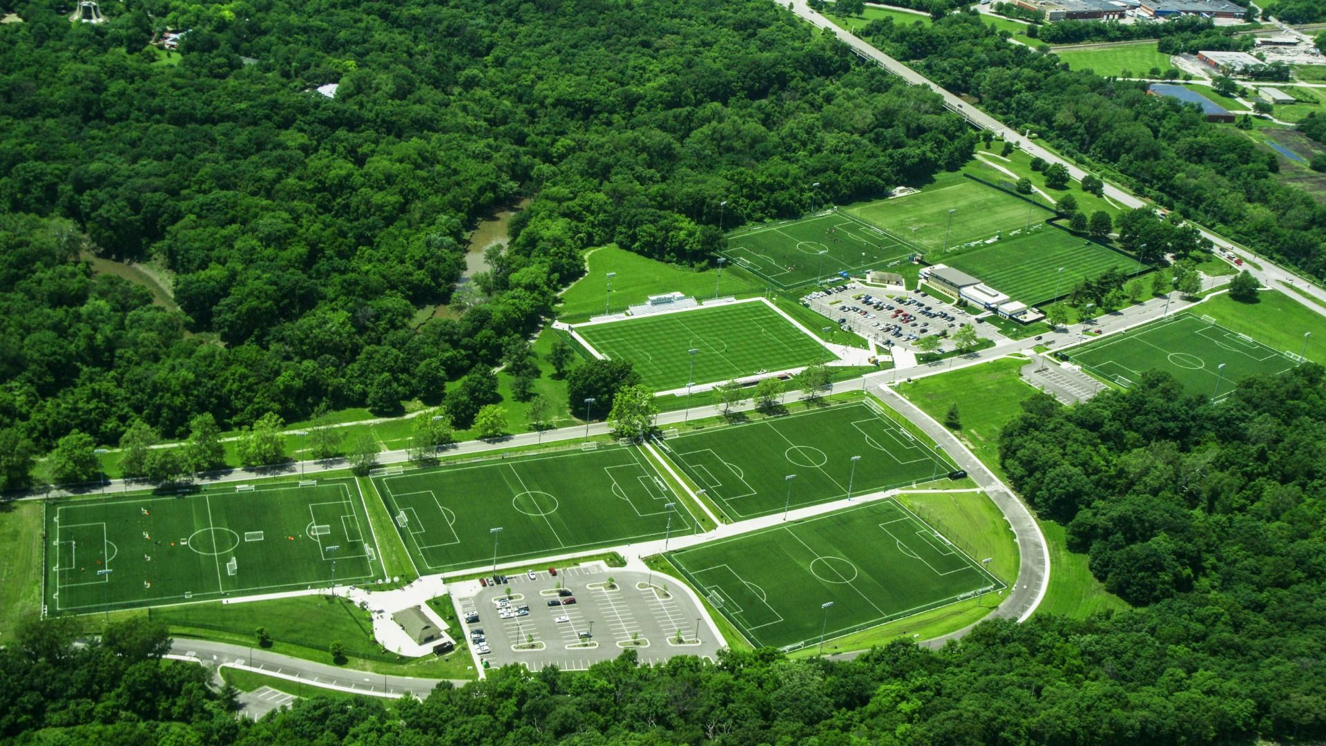 Aerial view of multiple lush green soccer fields at Swope Soccer Village surrounded by dense trees, parking lots with cars, and nearby roads under clear daylight.