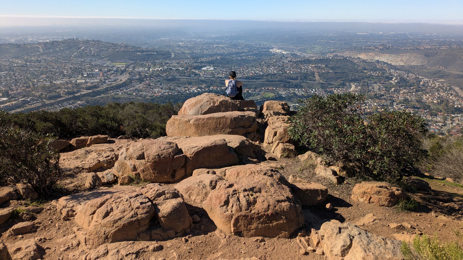 Person with a backpack sitting on large rocks atop a hill, overlooking a vast cityscape under clear blue sky with greenery and houses below.
